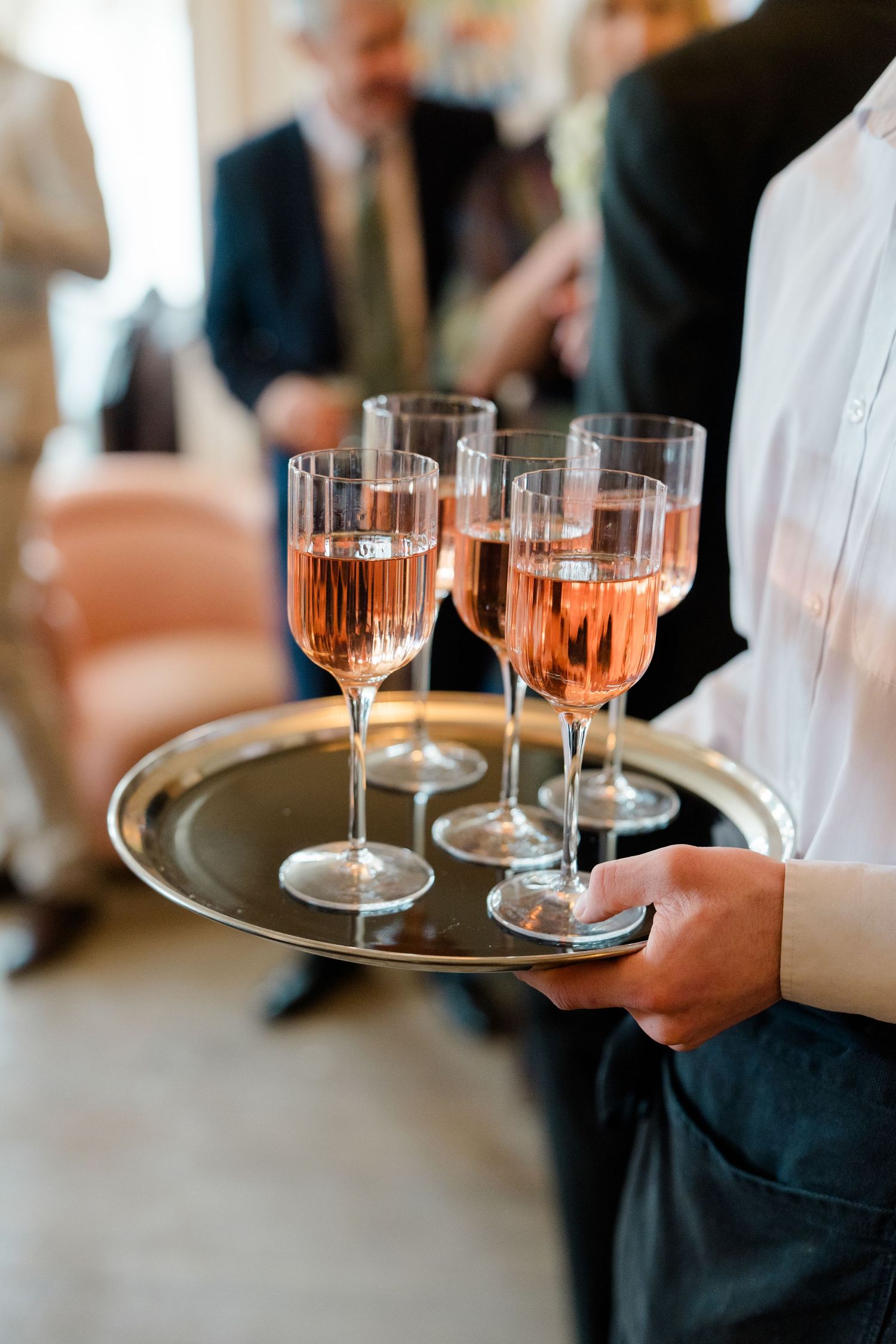 Waiter serving rose champagne in elegant flute glasses on a silver tray at a wedding reception.