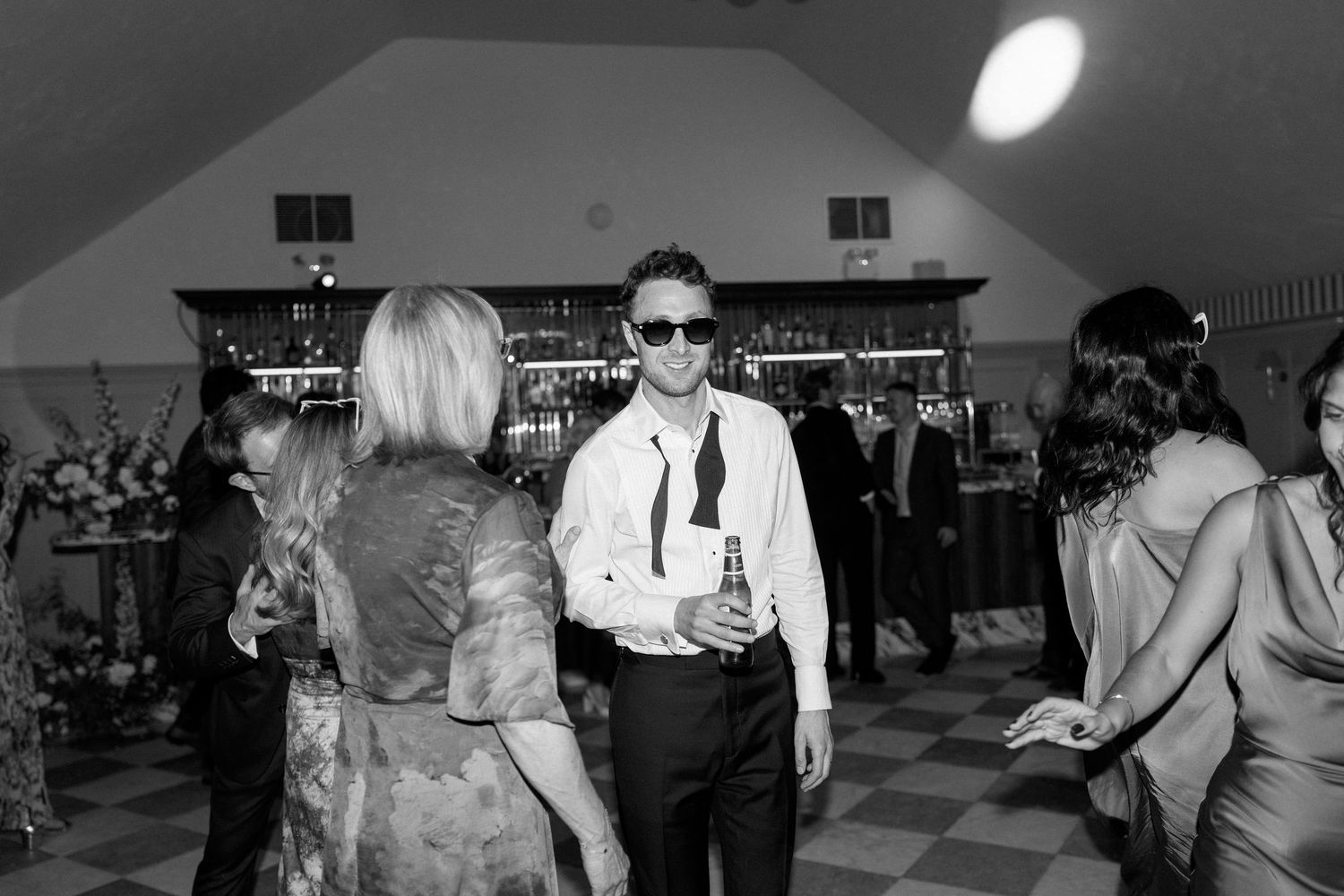 Wedding guest in white shirt and sunglasses dancing at lively reception near bar in black and white.