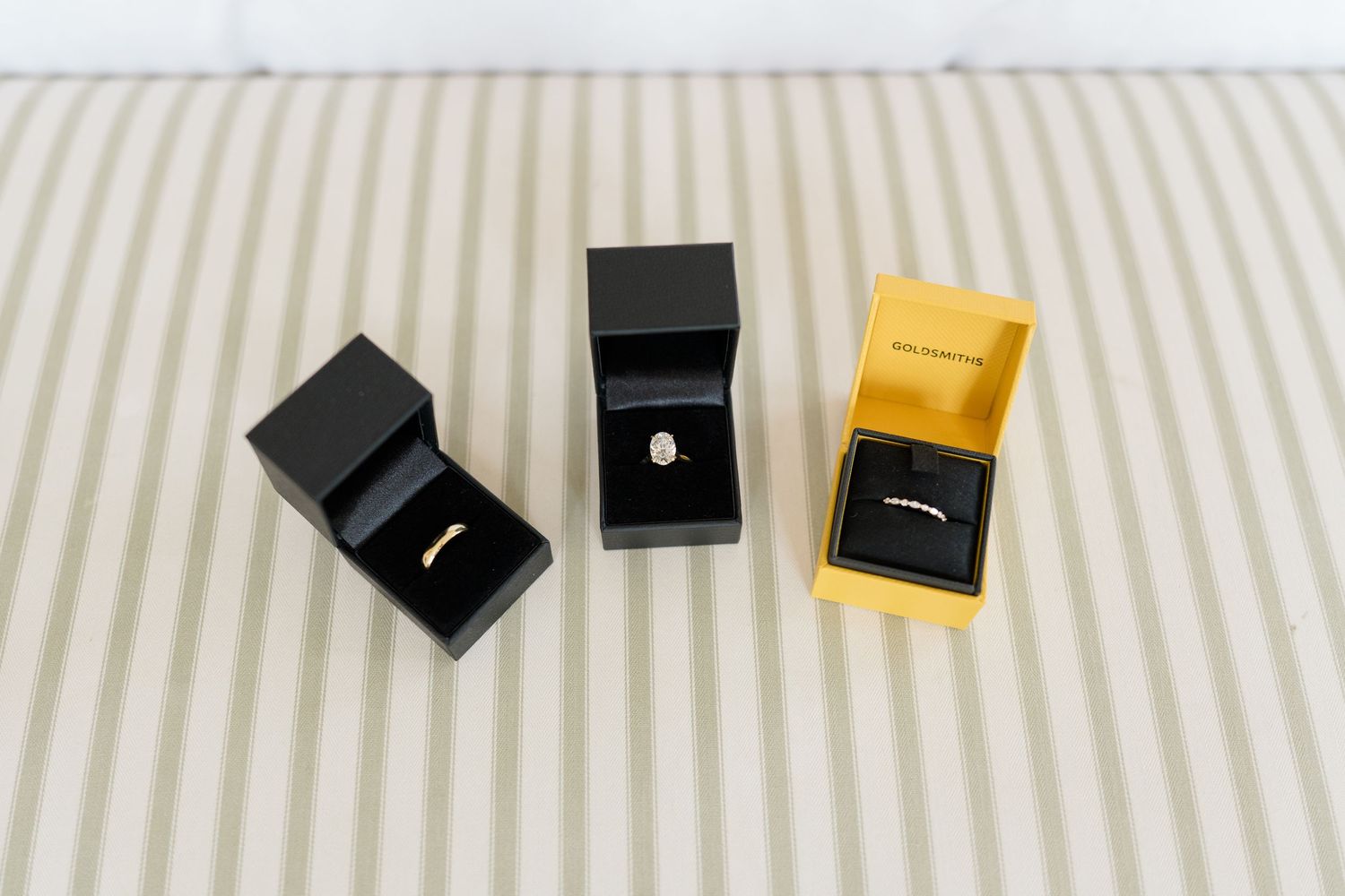 Three wedding rings displayed in open black and yellow jewelry boxes on striped surface.
