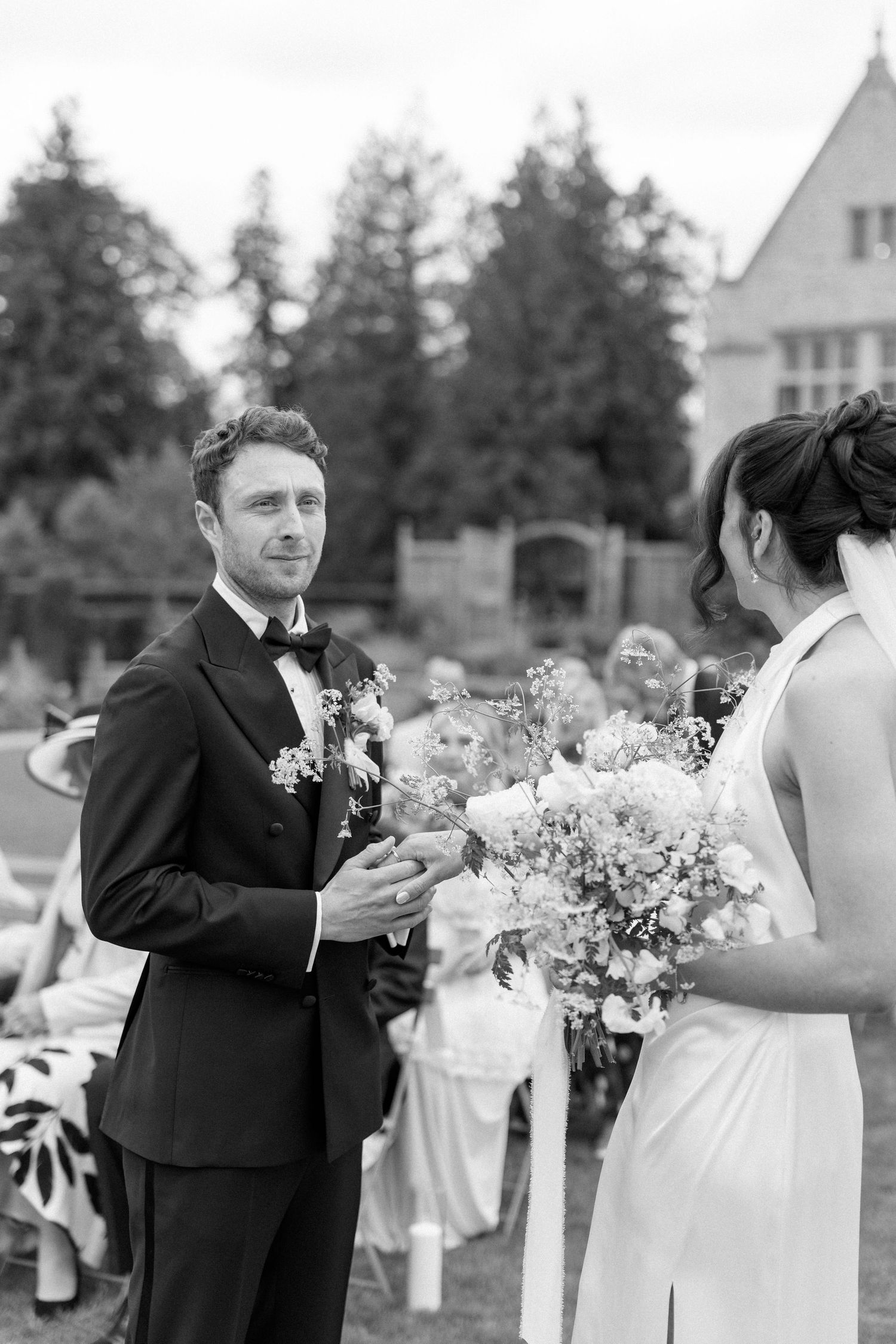 Groom in black tuxedo exchanges vows with bride holding floral bouquet during outdoor wedding ceremony.