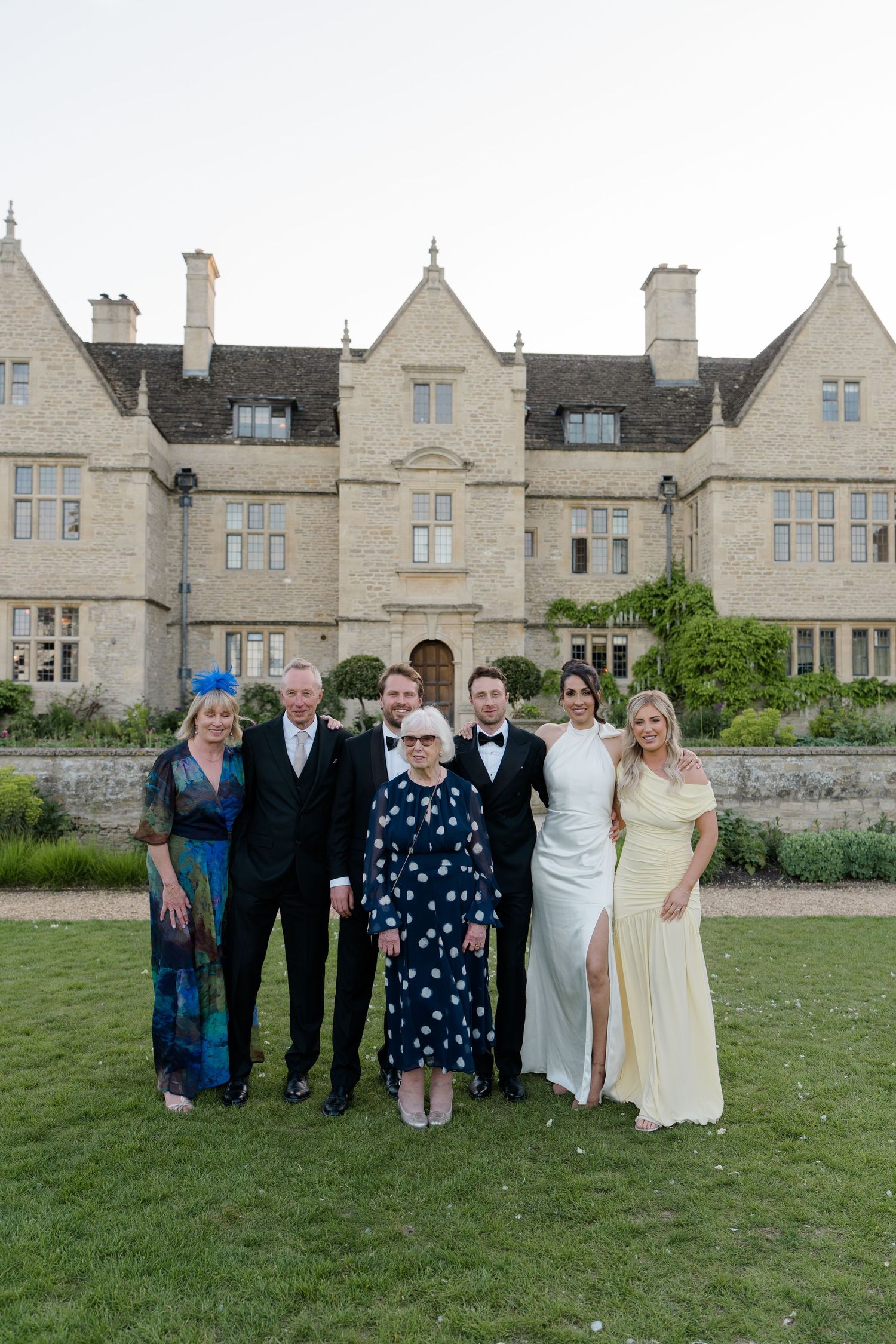 Wedding party group photo of six people posing in front of a grand English manor house at dusk.