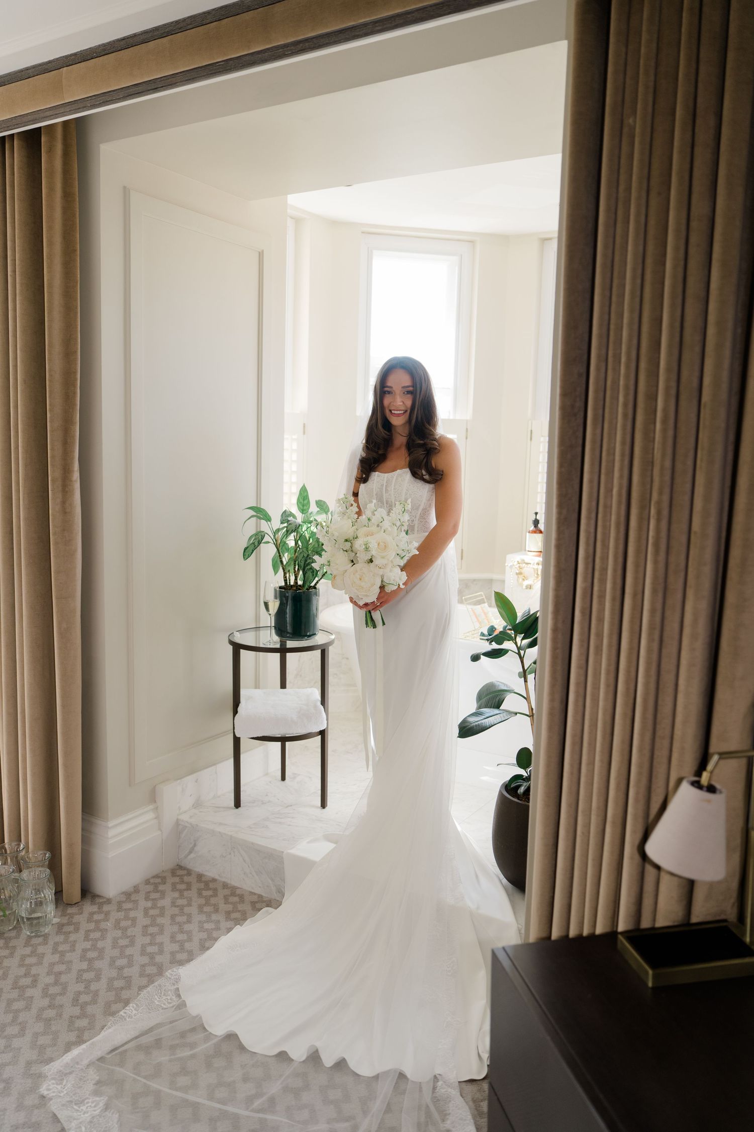 Full-length portrait of smiling bride in fitted mermaid wedding dress with train holding white floral bouquet.