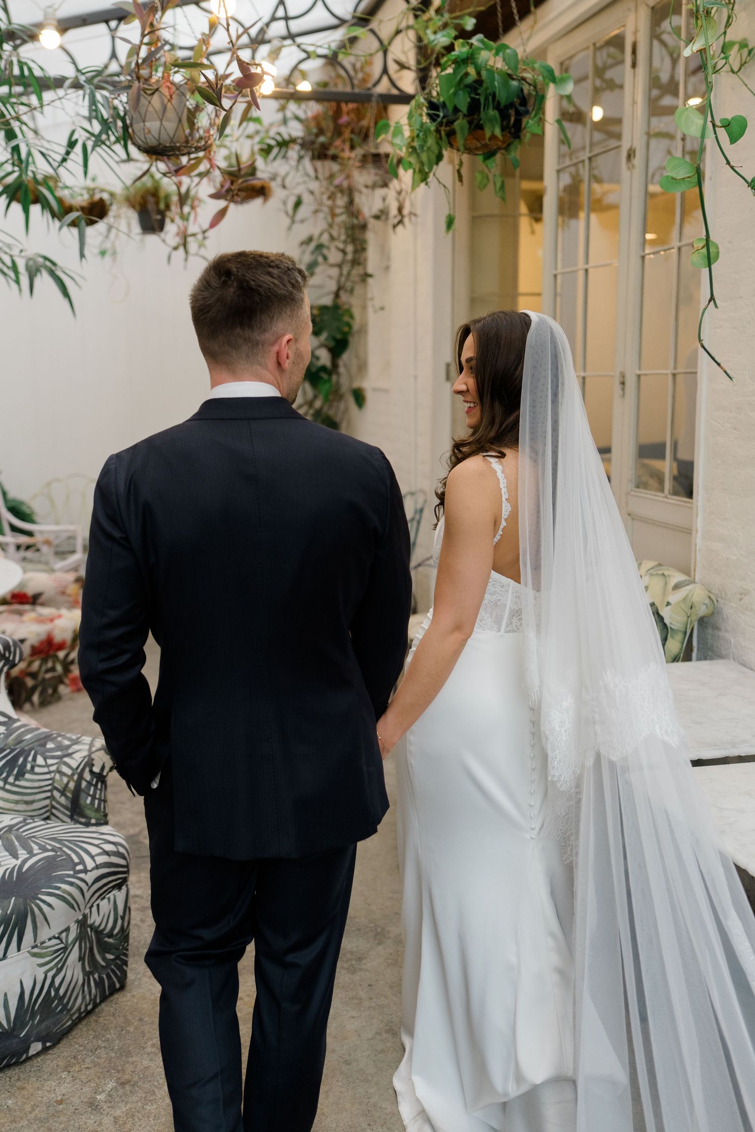 Bride in white gown and veil holds hands with groom in black suit walking through lush indoor garden venue.