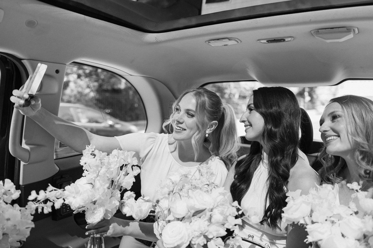 Three bridesmaids in white dresses laugh and take selfies in a car with white floral bouquets.
