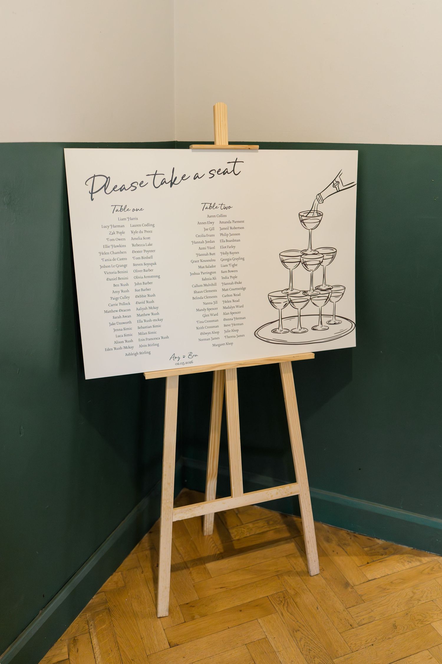 Wedding seating chart on easel with champagne tower illustration reads Please Take a Seat.
