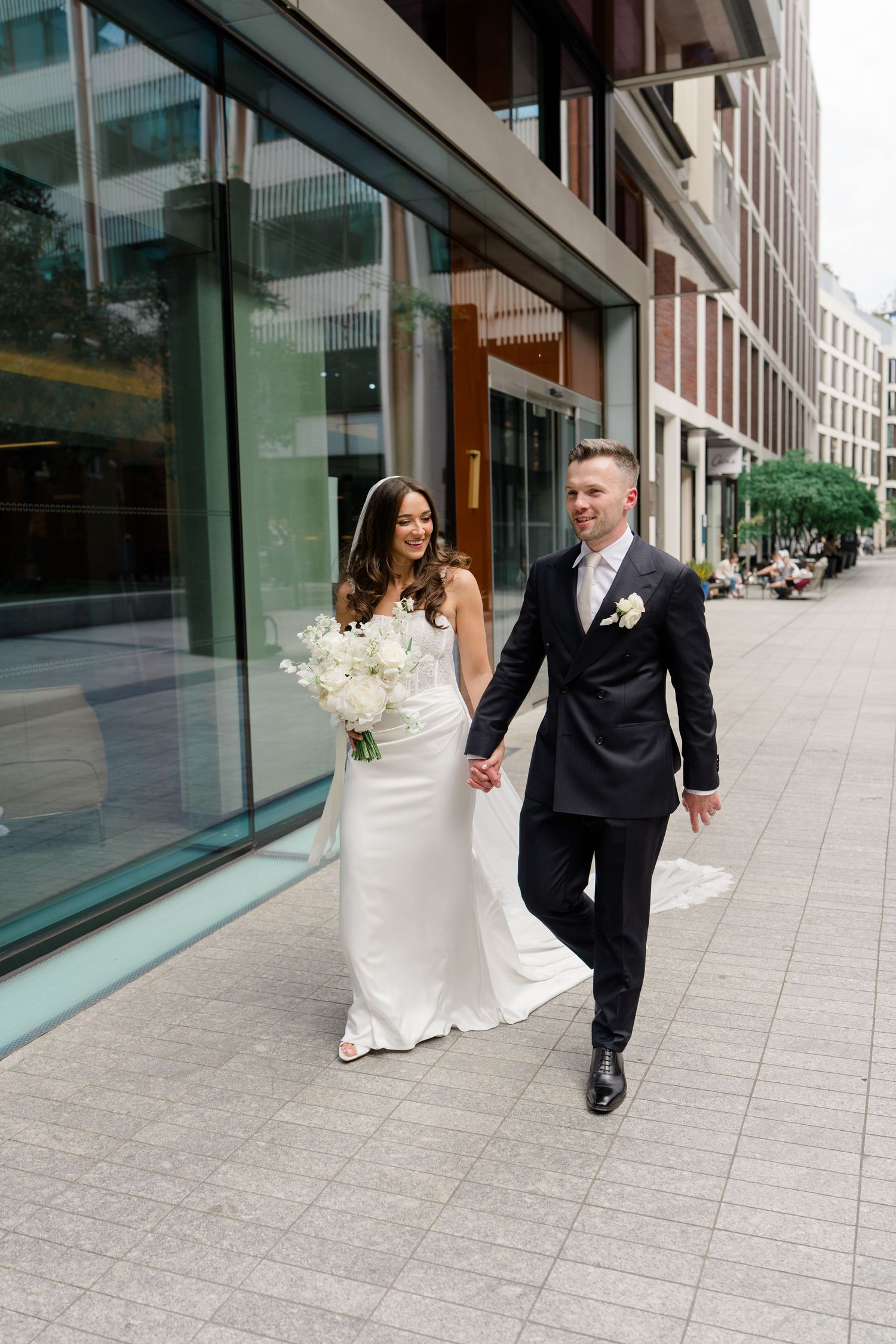 Bride and groom walking hand in hand outside a modern glass building, bride in white satin gown holding white bouquet.
