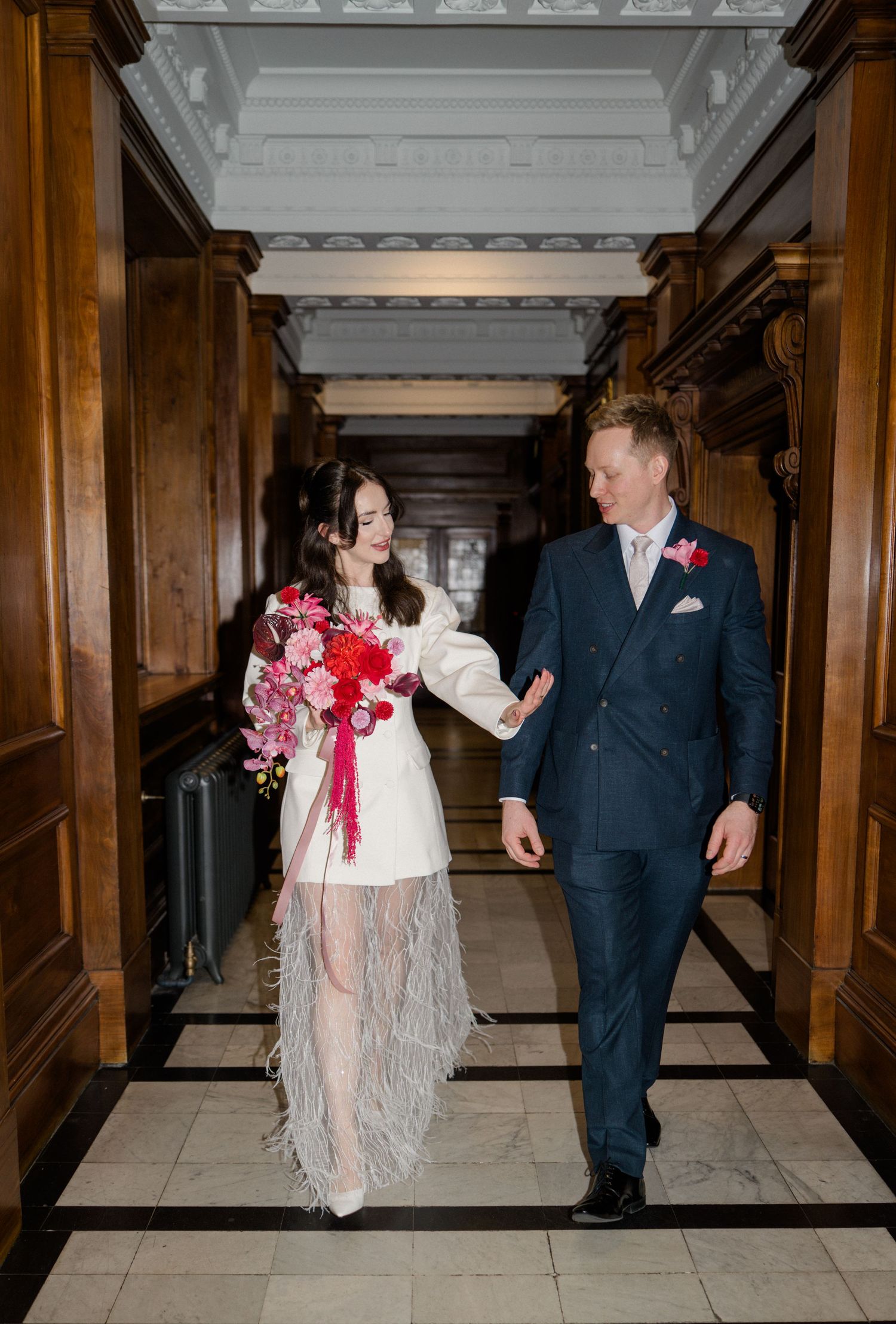 Bride in white fringe mini dress with red bouquet walks with groom in navy suit.