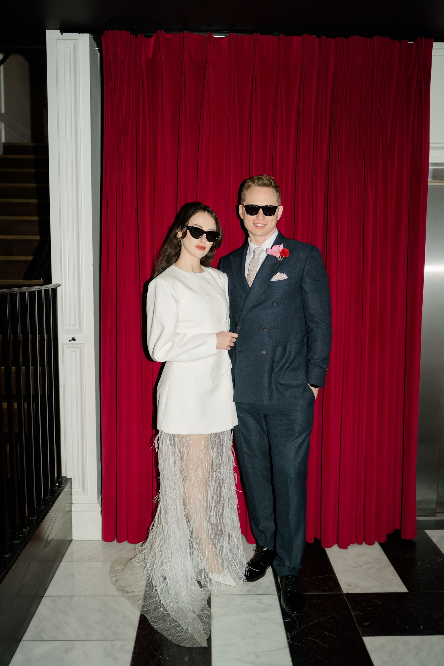 Stylish bride and groom pose in sunglasses before a red velvet curtain, bride in feathered white mini dress.
