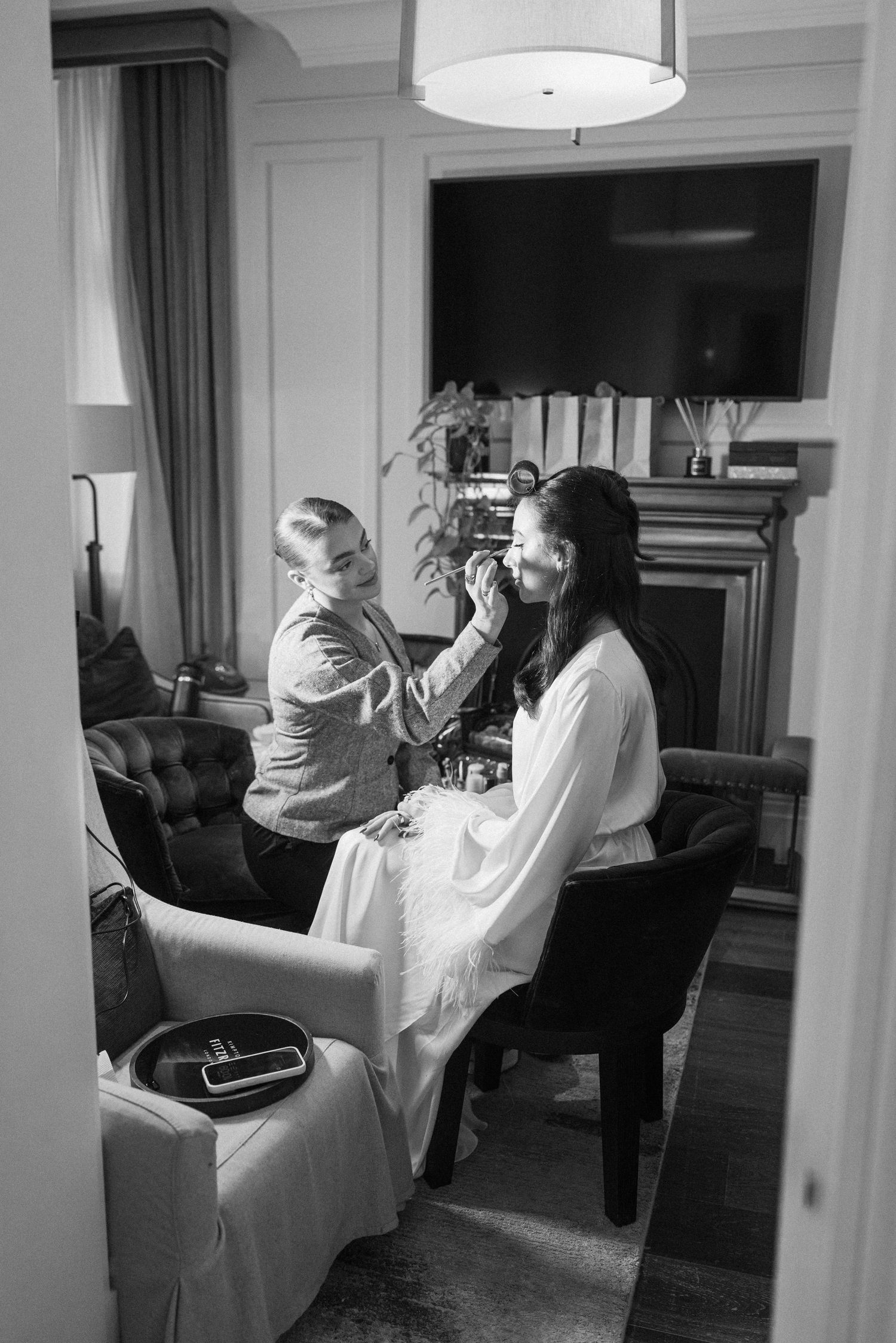 Black and white photo of bride getting makeup applied by artist in elegant hotel suite.