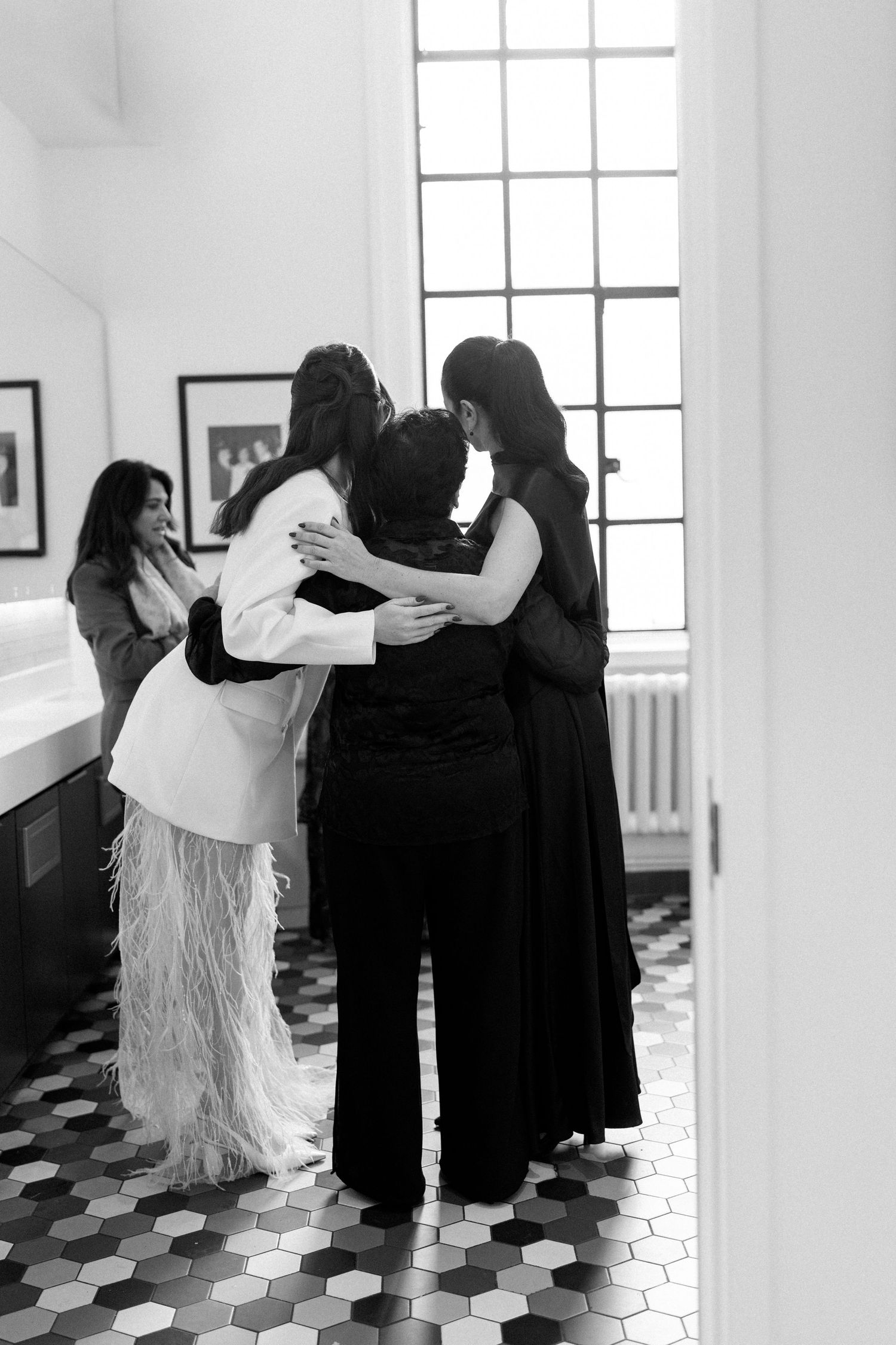 Black and white photo of bride in feathered white suit embracing two guests in bright bathroom.