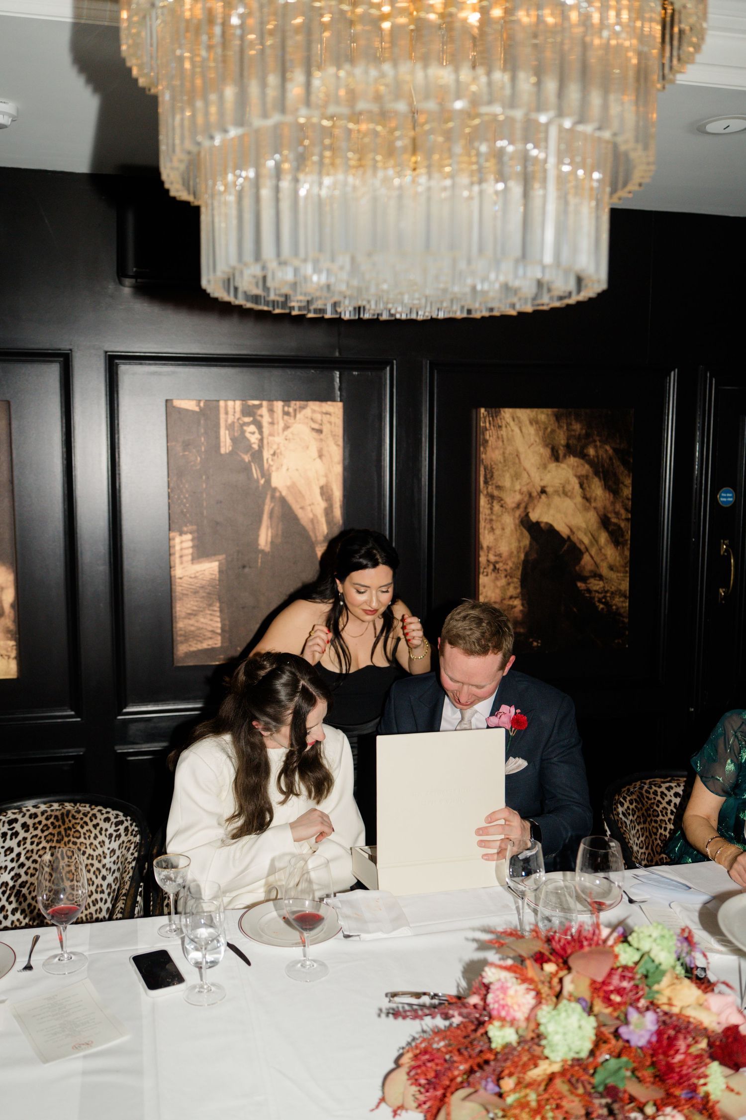 Bride and groom reviewing menu at wedding reception under gold chandelier with colorful floral centerpiece.