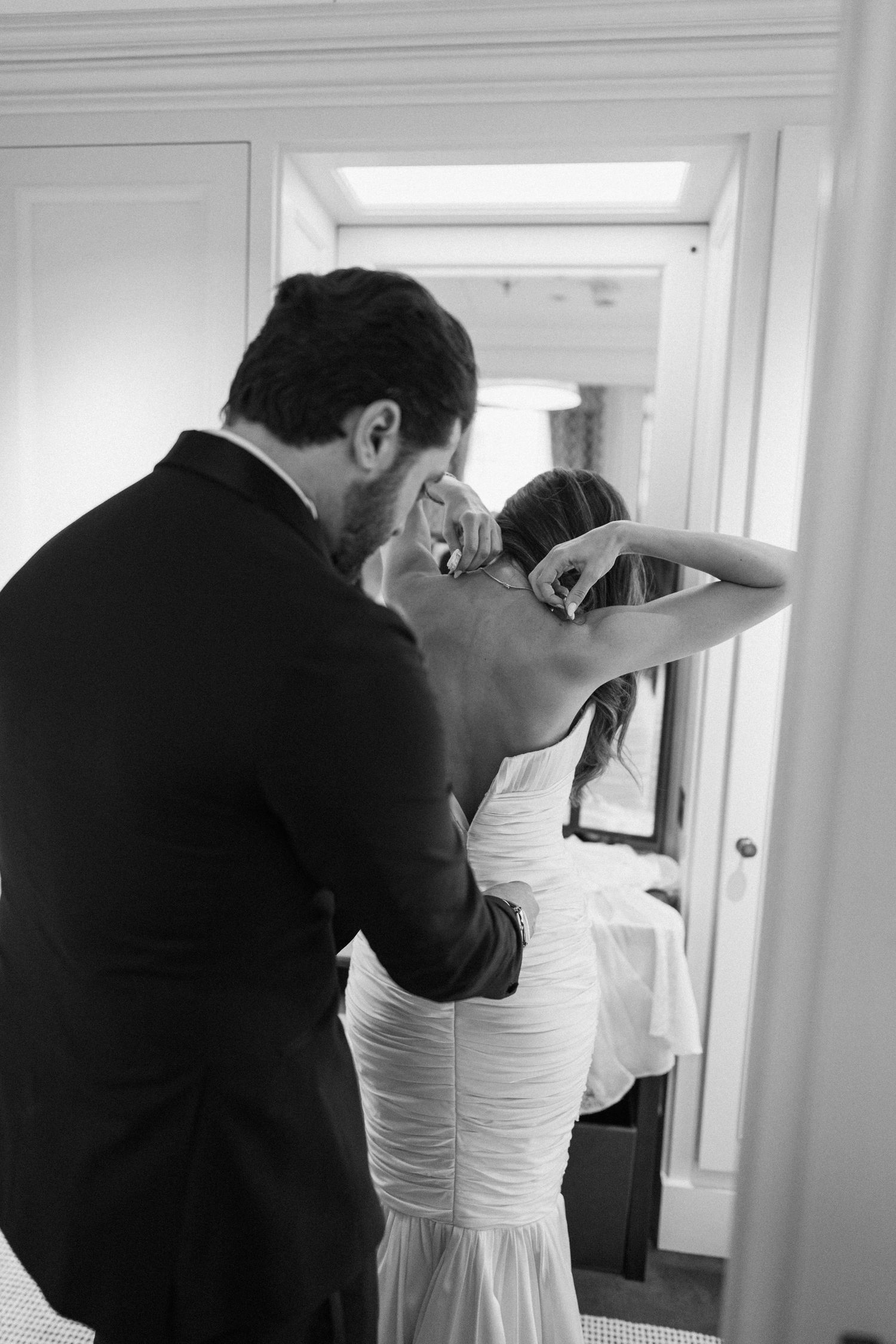 Black and white photo of groom helping bride with her dress before wedding ceremony.