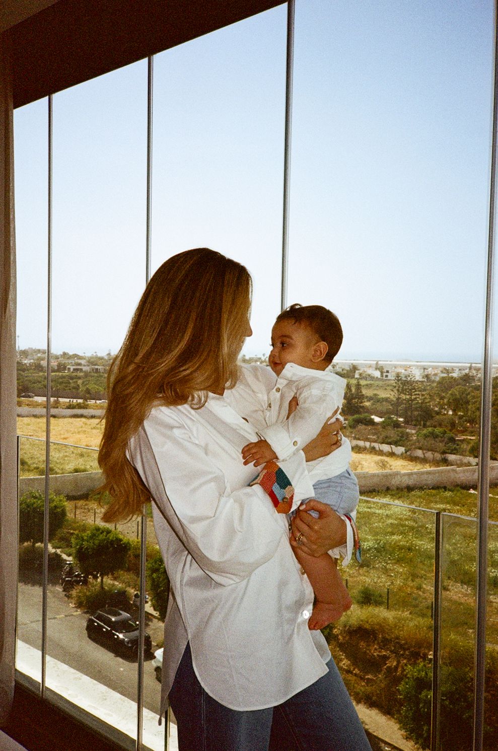 A woman with long blonde hair in a white shirt holds a baby near large windows with a scenic coastal view.