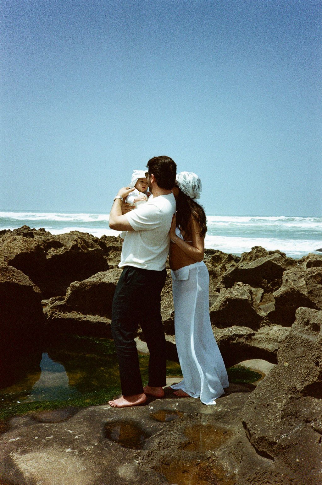 Couple holding baby kissing on rocky beach with ocean waves and clear blue sky in background.