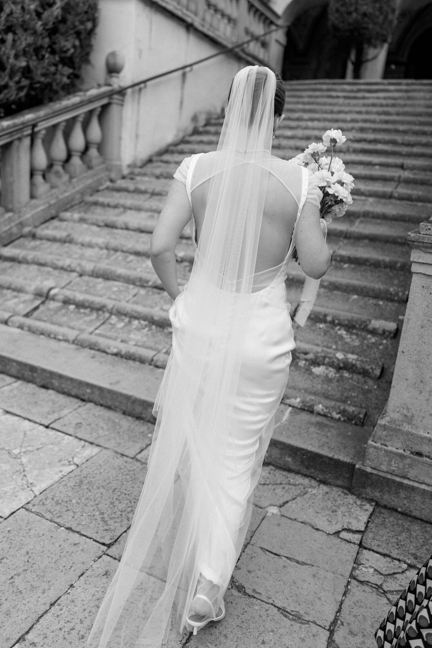 Black and white photo of bride in flowing veil ascending stone steps.