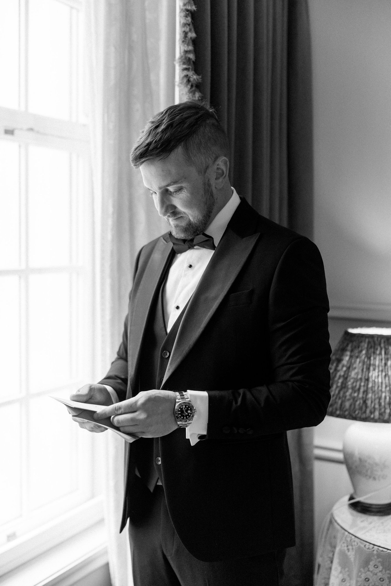 Person in a black tuxedo checking their phone near a bright window.