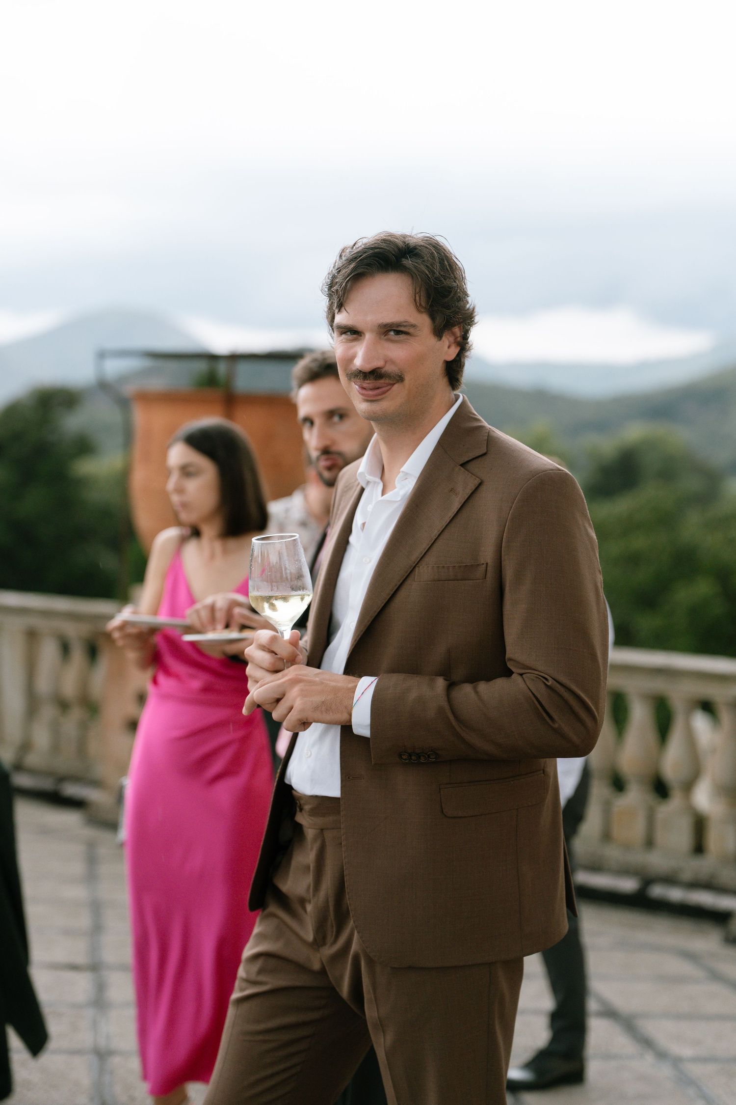 A guest in a brown suit holding a wine glass on a terrace with mountain views.