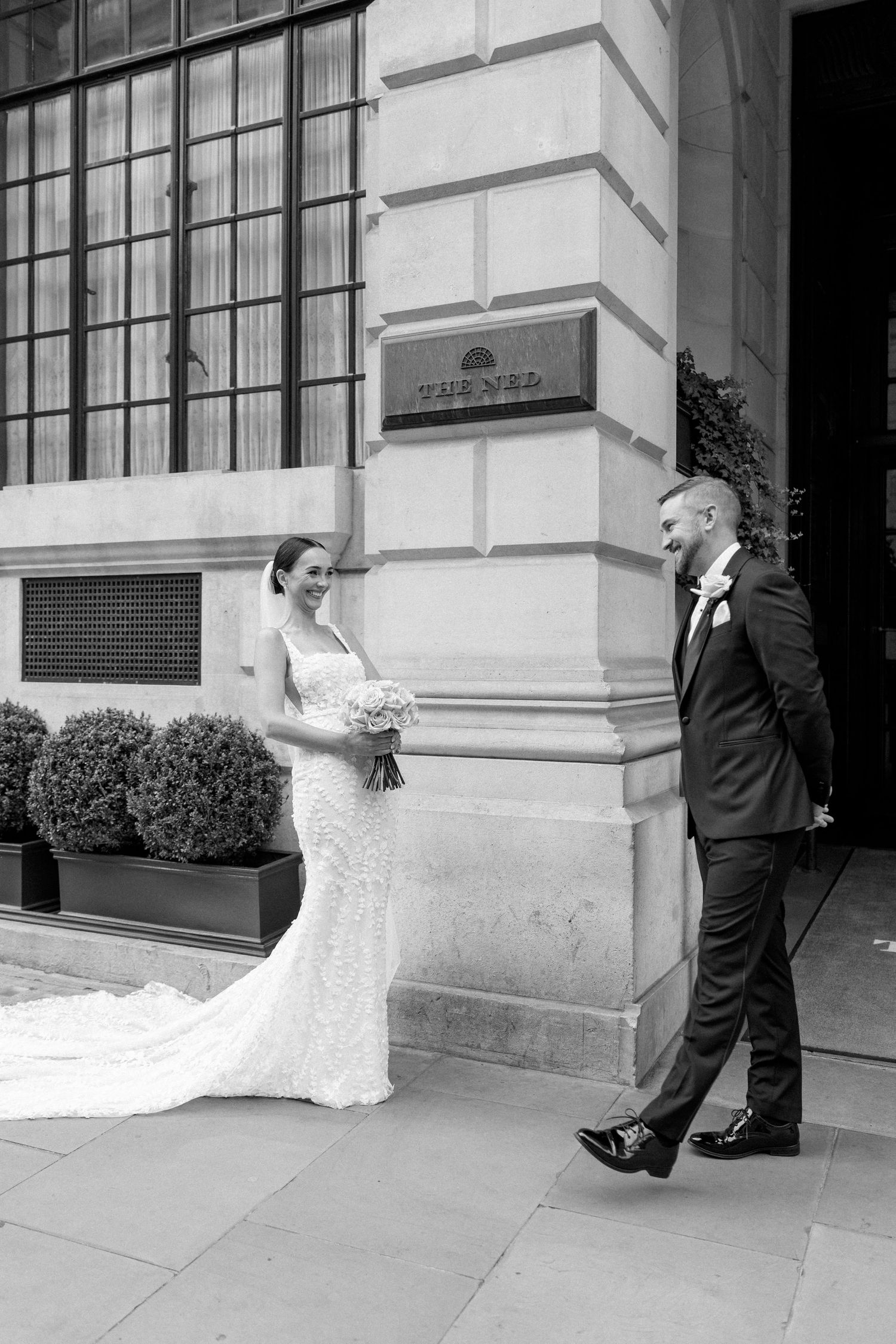 A black and white wedding photo showing a couple in formal attire outside an elegant building entrance.
