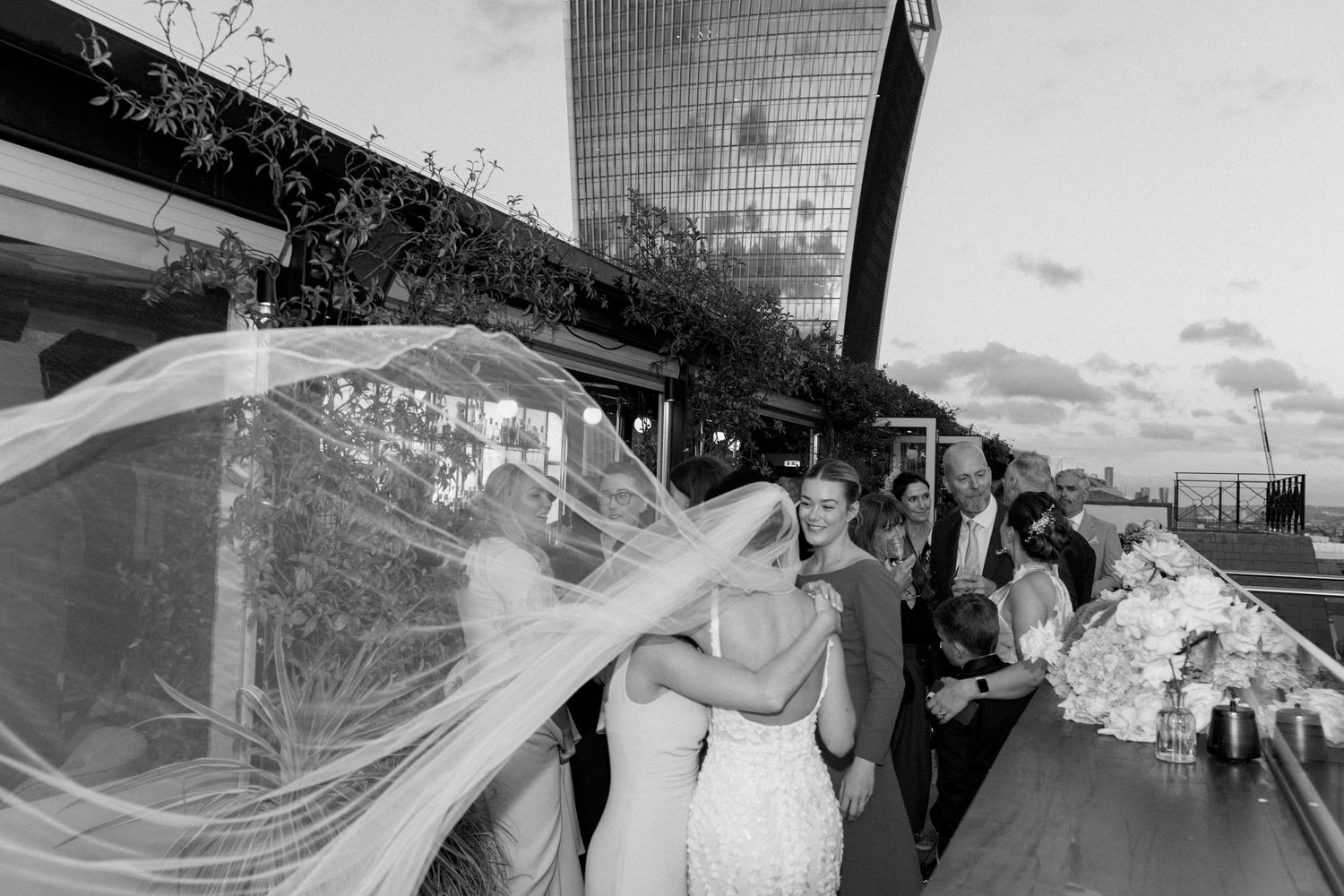 Wedding guests celebrate on an outdoor terrace with a modern building in the background during sunset.