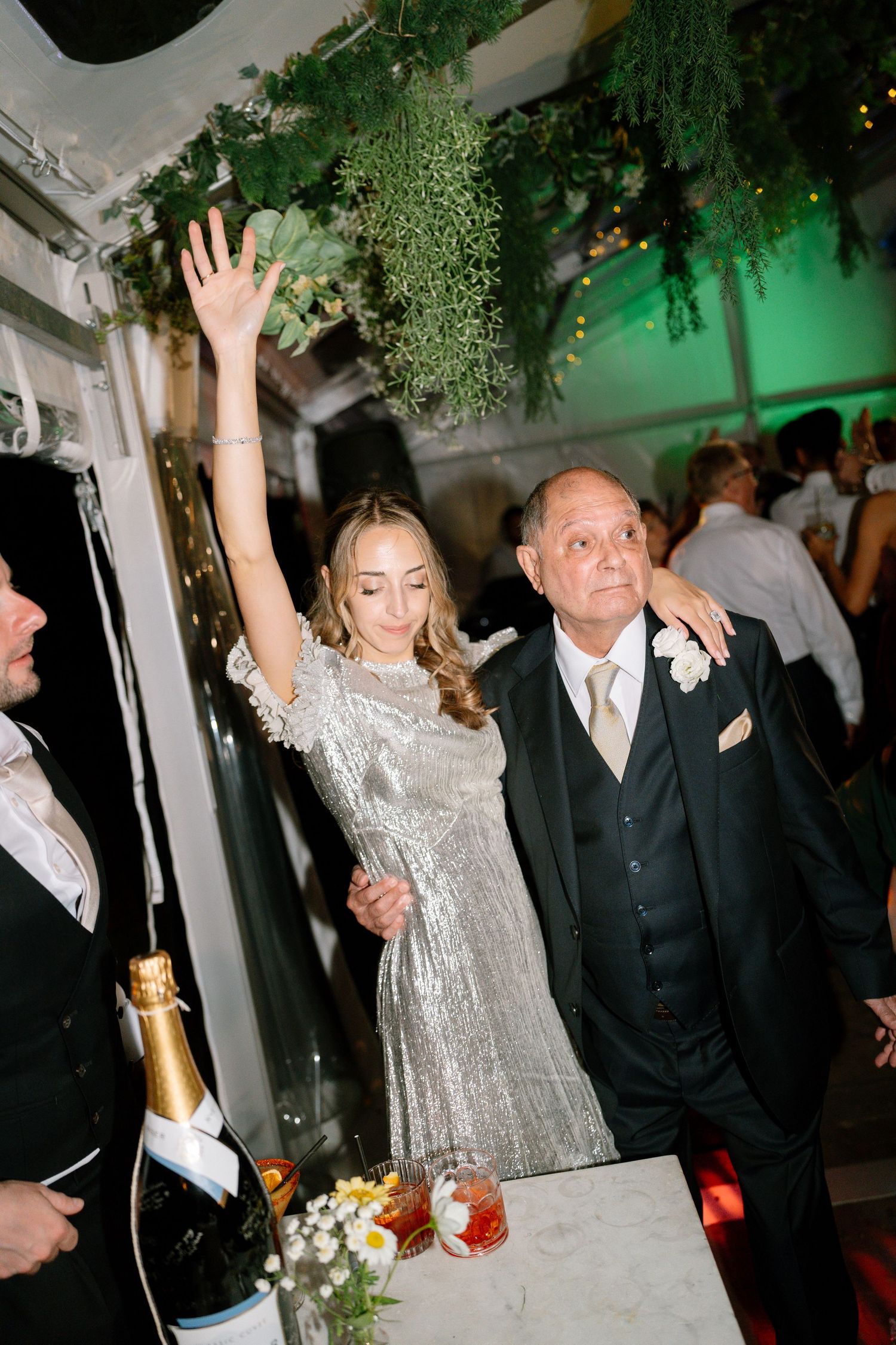 Party guests celebrate with raised arms under hanging greenery at an evening event.