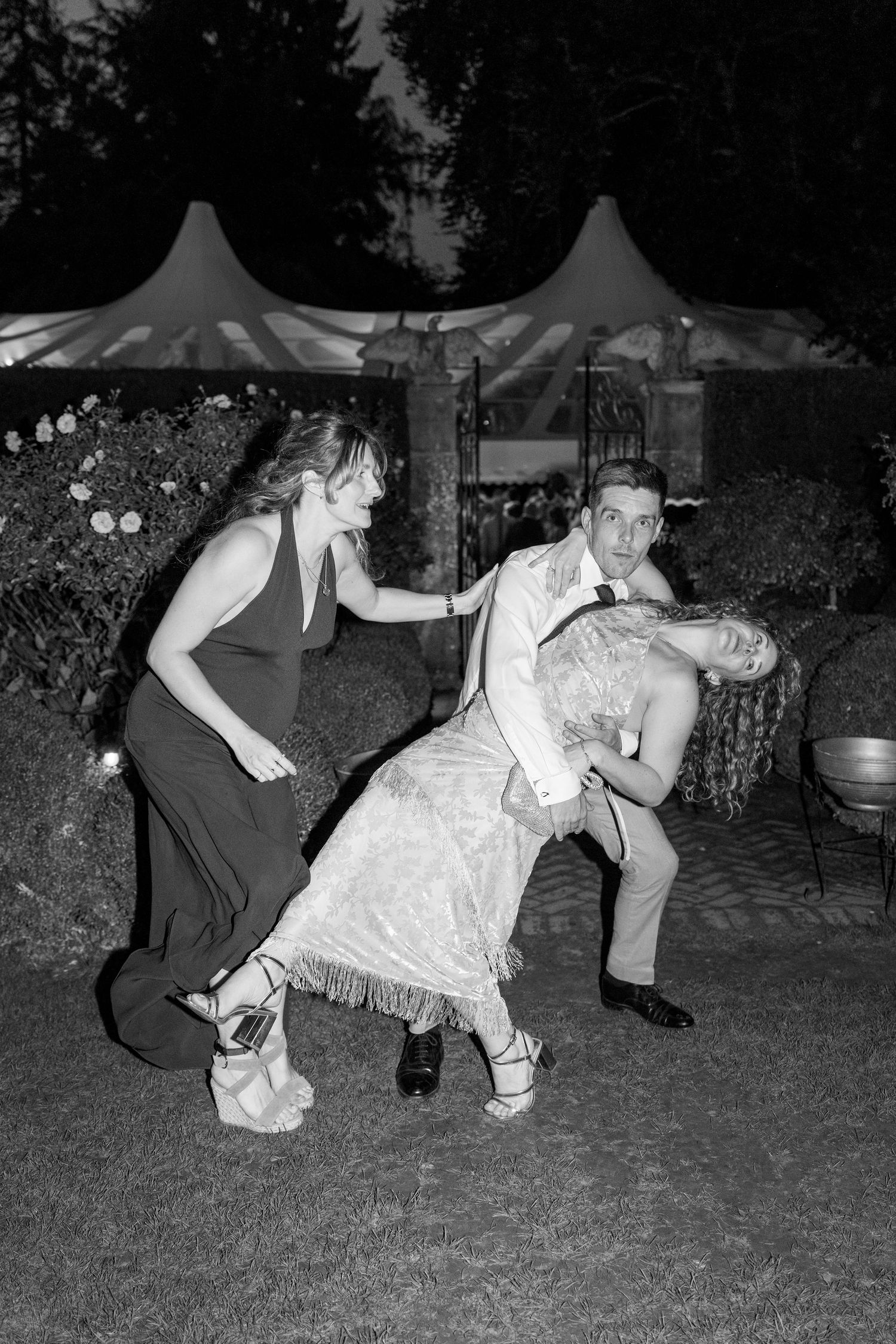 Wedding guests dance and pose playfully at an outdoor reception under white tent canopies.