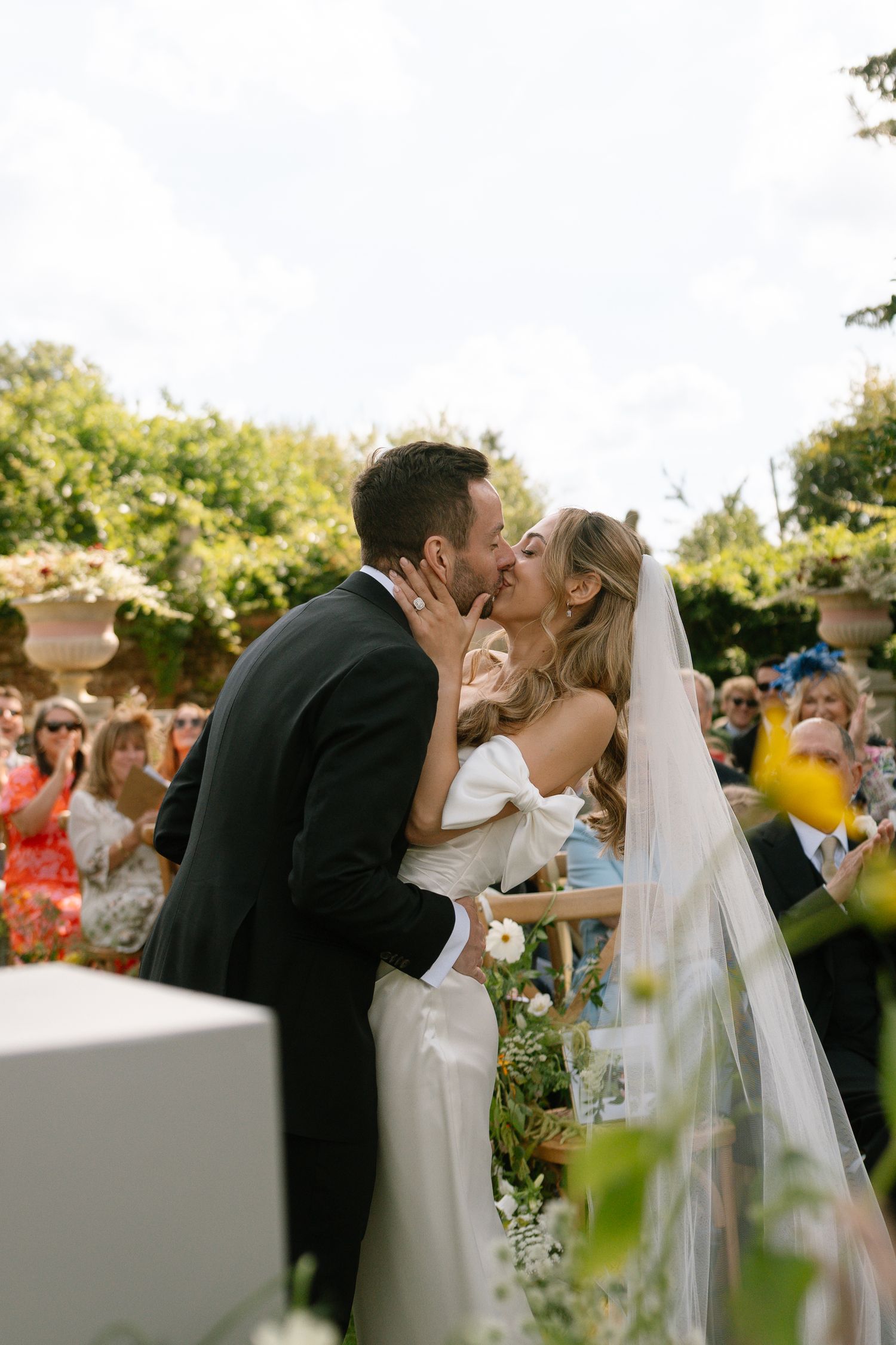 Wedding couple embraces during their first kiss at a garden ceremony with guests watching.