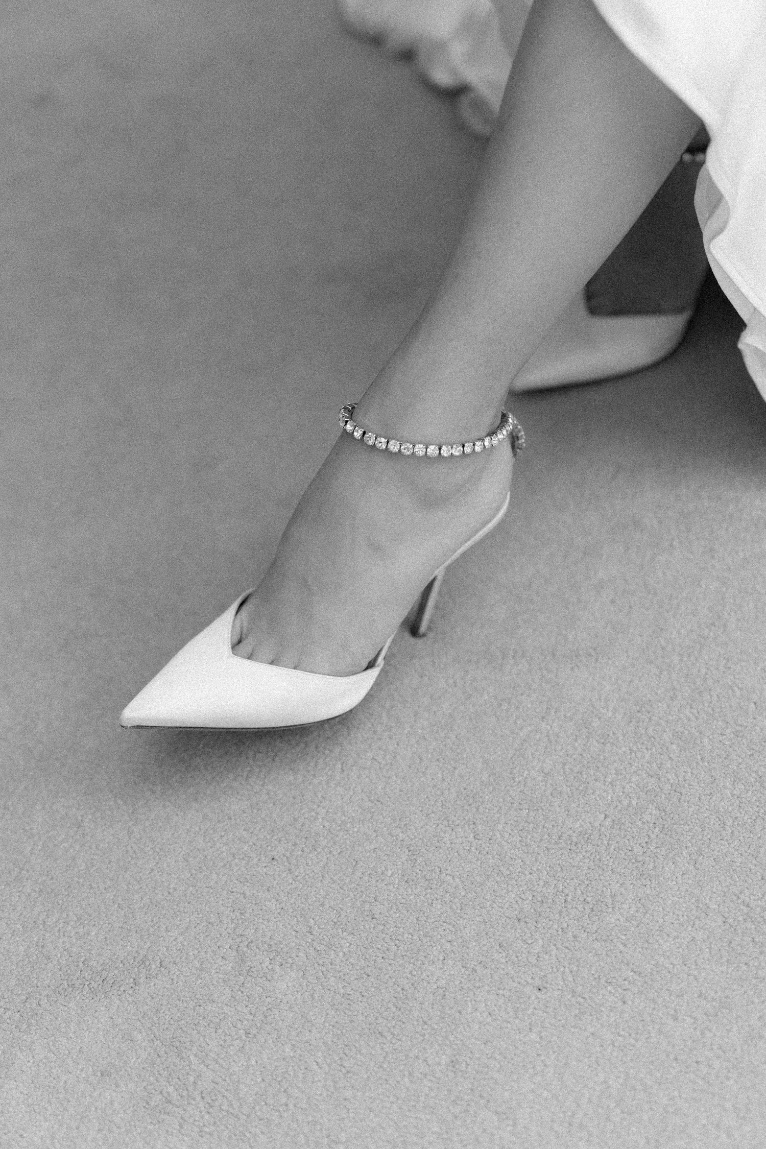 Close-up detail of anklet and white dress hem against grey carpet.