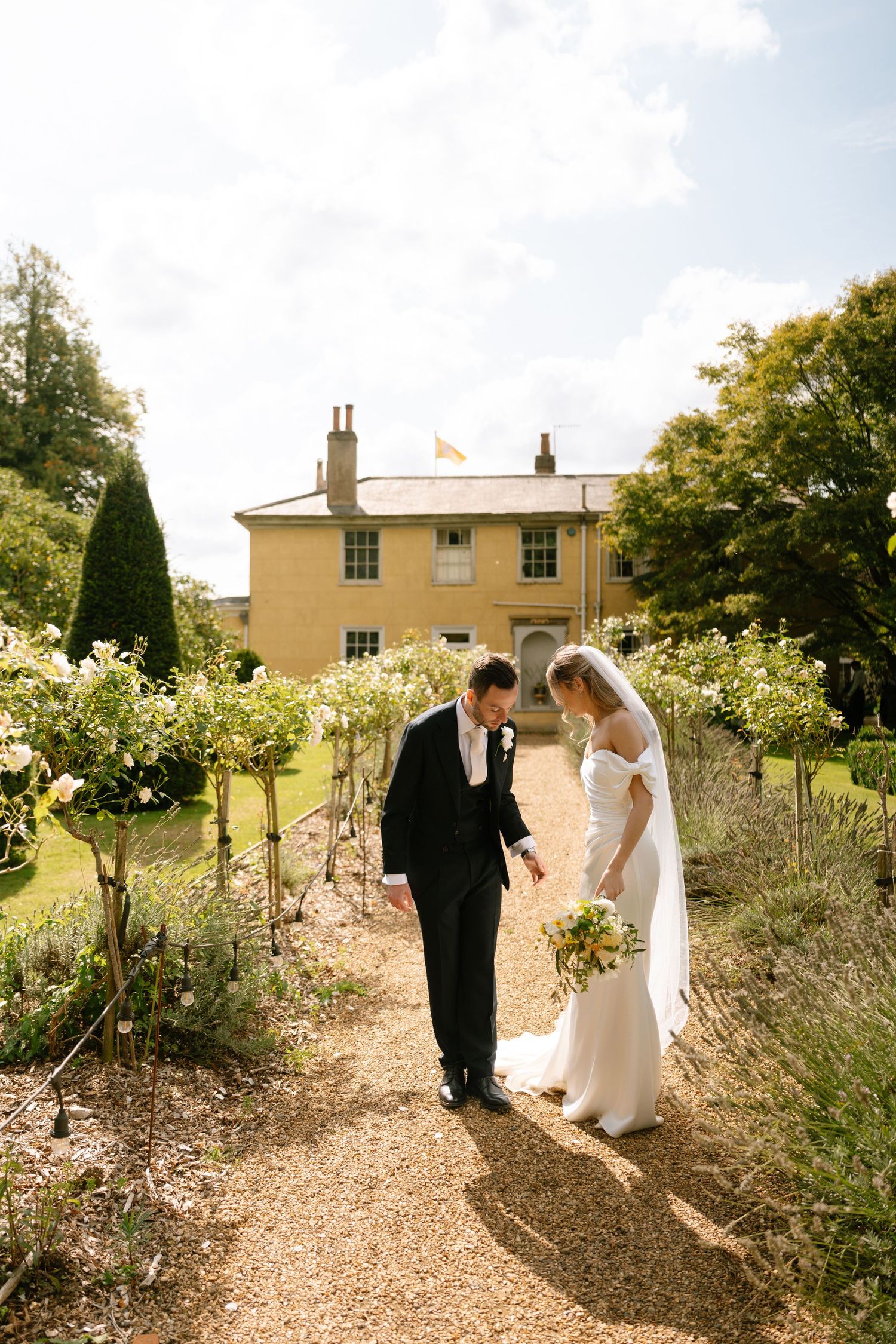 A wedding couple walks along a garden path lined with lavender and roses near a yellow stone manor house.