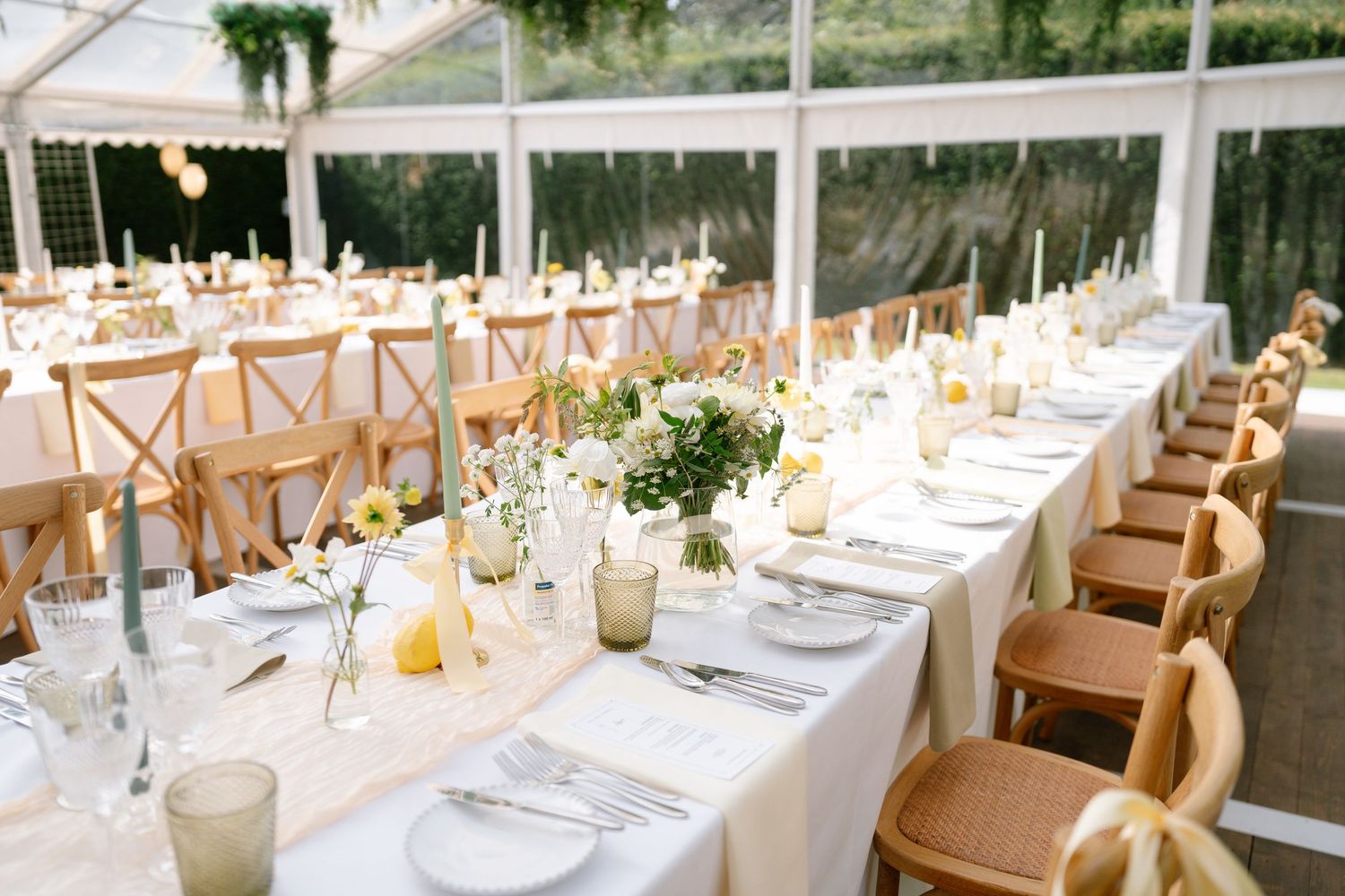Elegant wedding reception setup with white tablecloths, wooden chairs, and hanging greenery in a clear tent.