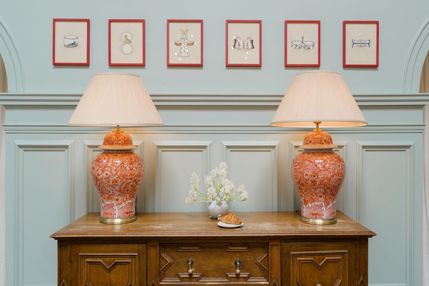 Elegant wooden sideboard with two orange ceramic ginger jar lamps and red-framed botanical prints on a pale blue wall.