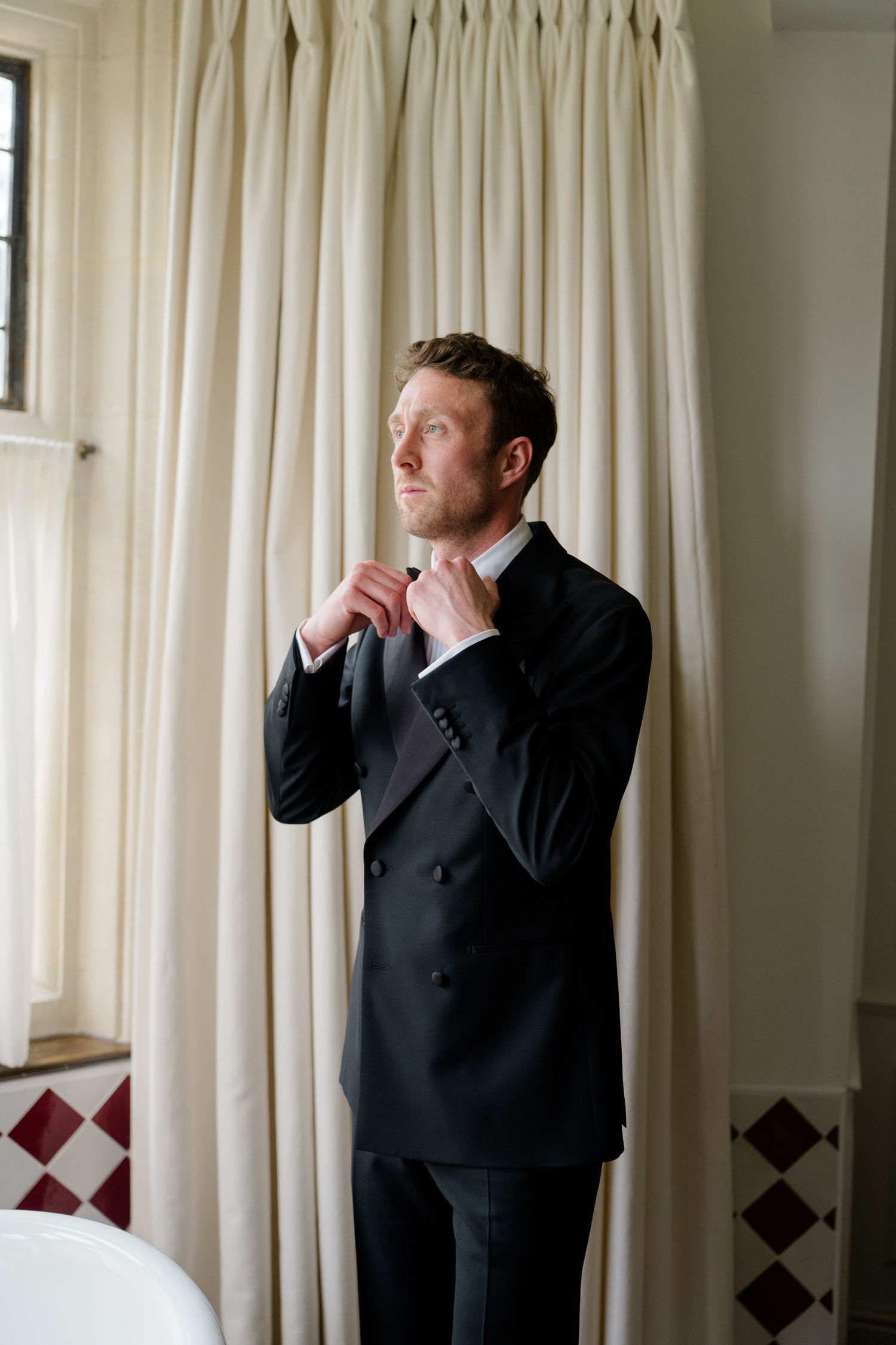 A groom in a black tuxedo adjusting his tie in front of cream curtains.