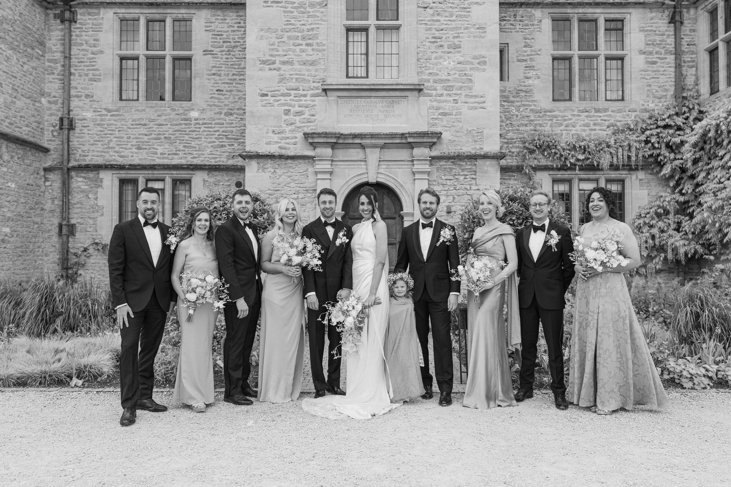 Black and white photo of full wedding party in formal attire posing outside a historic stone manor house.