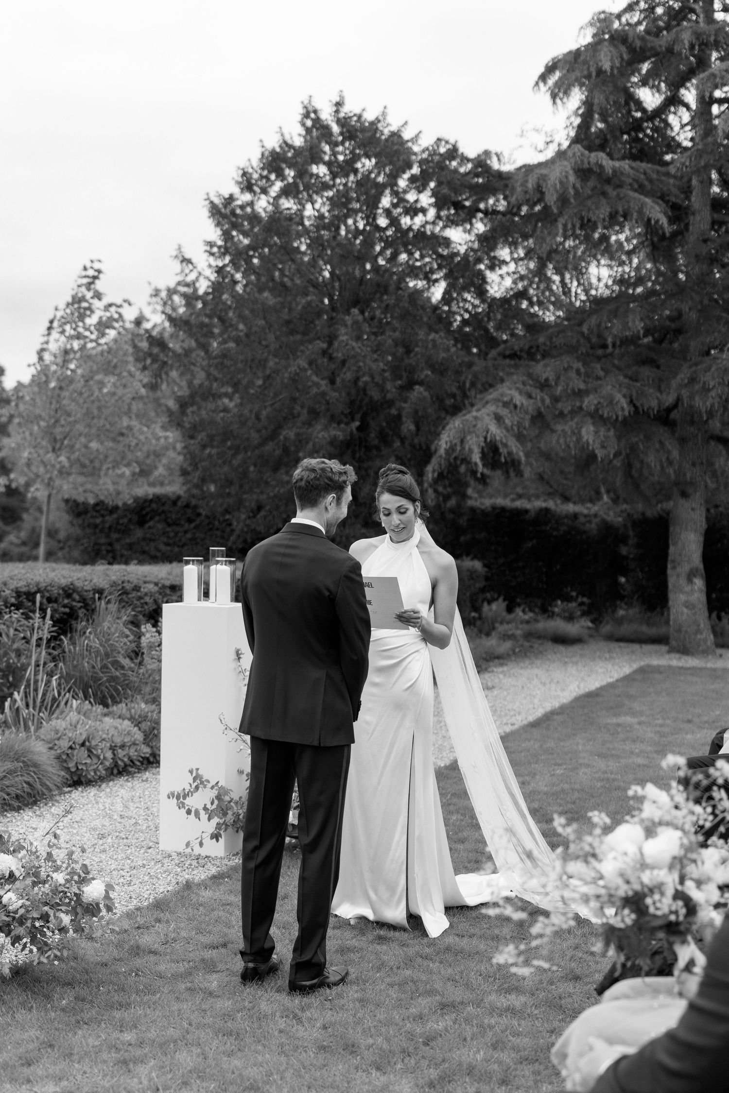 Bride reads wedding vows to groom during elegant outdoor garden ceremony in black and white.