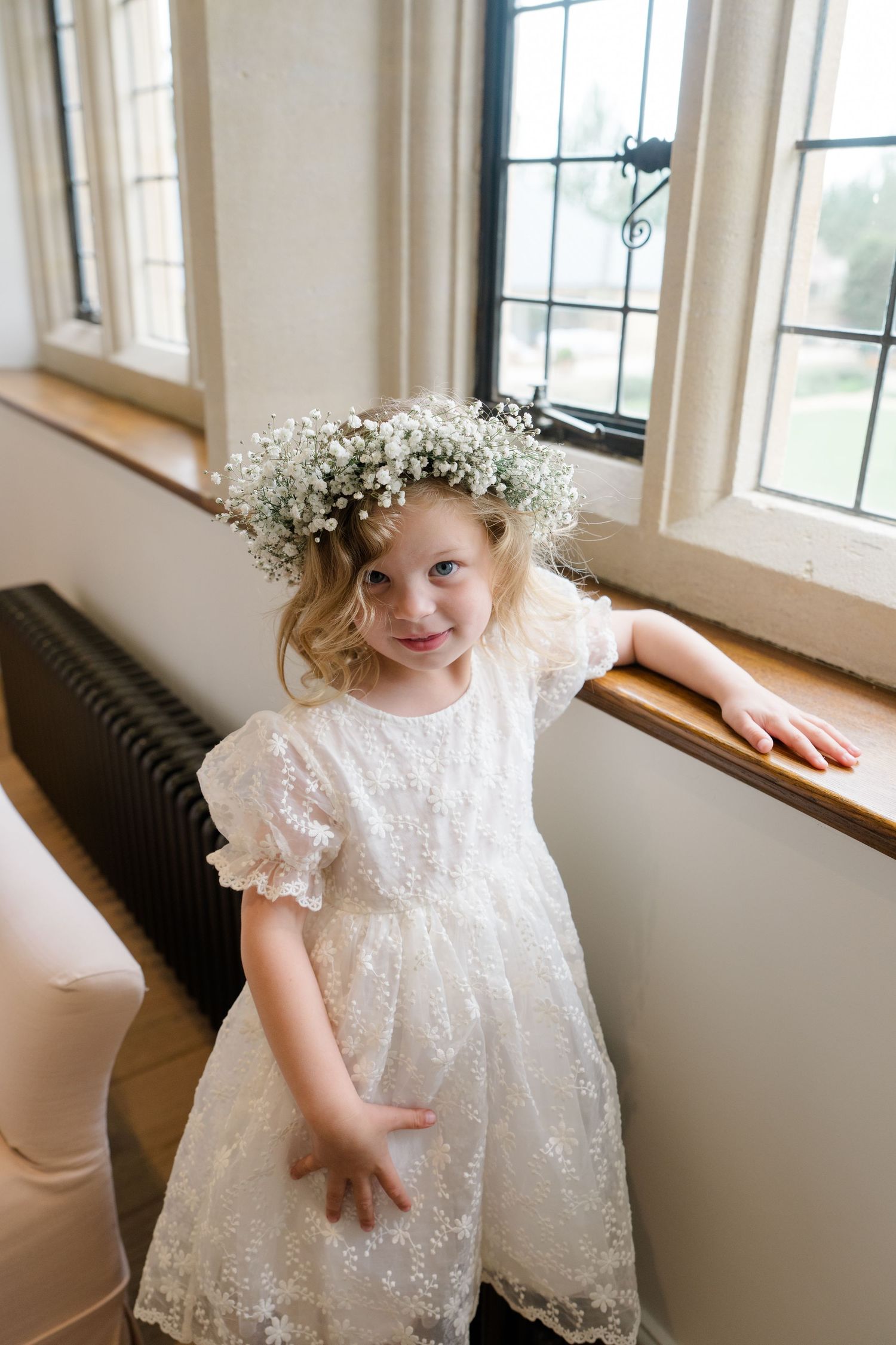 Adorable flower girl in white lace dress wearing baby's breath floral crown by window.