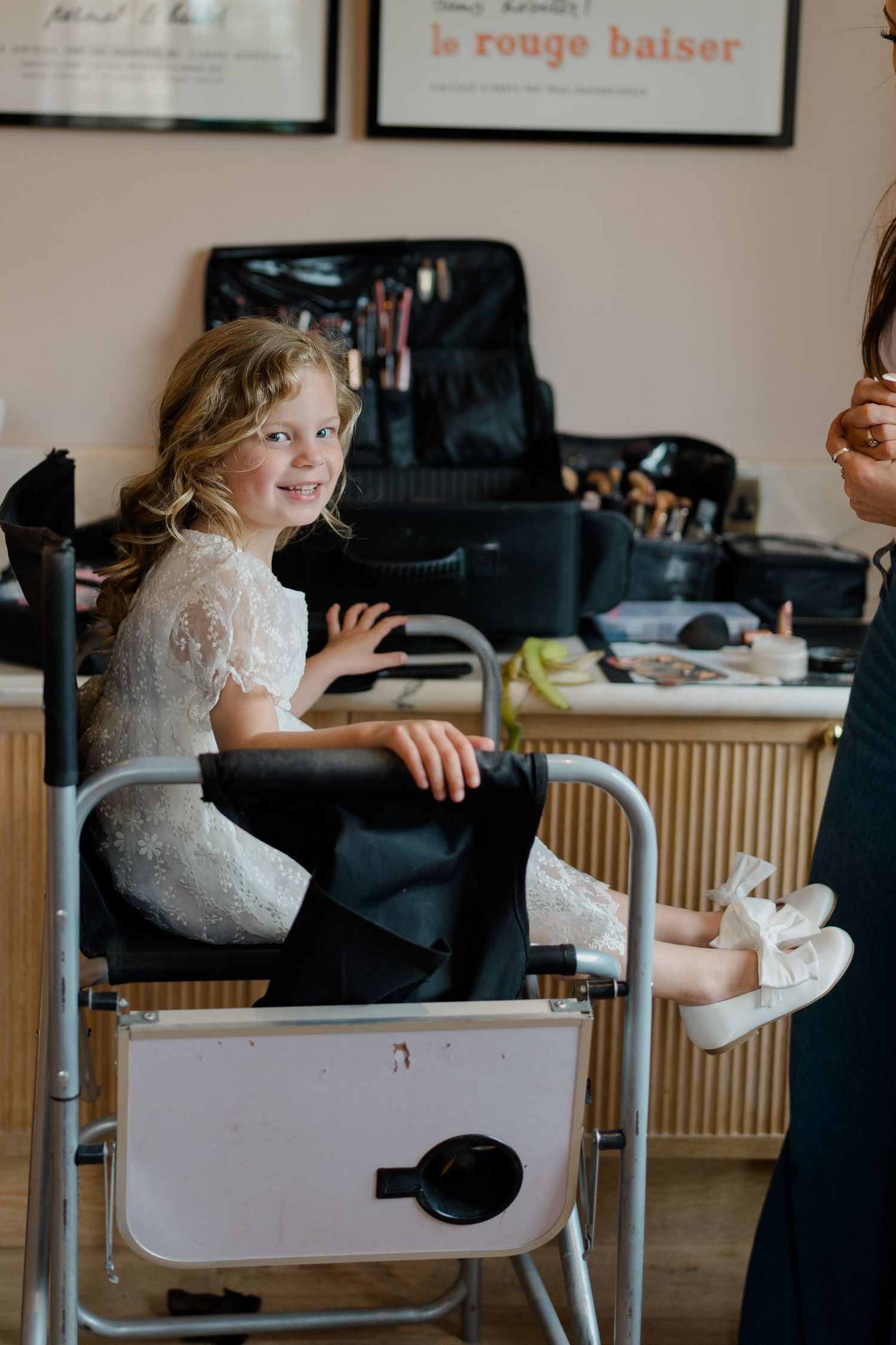 Smiling young flower girl in white lace dress sitting in a makeup chair at a bridal suite.