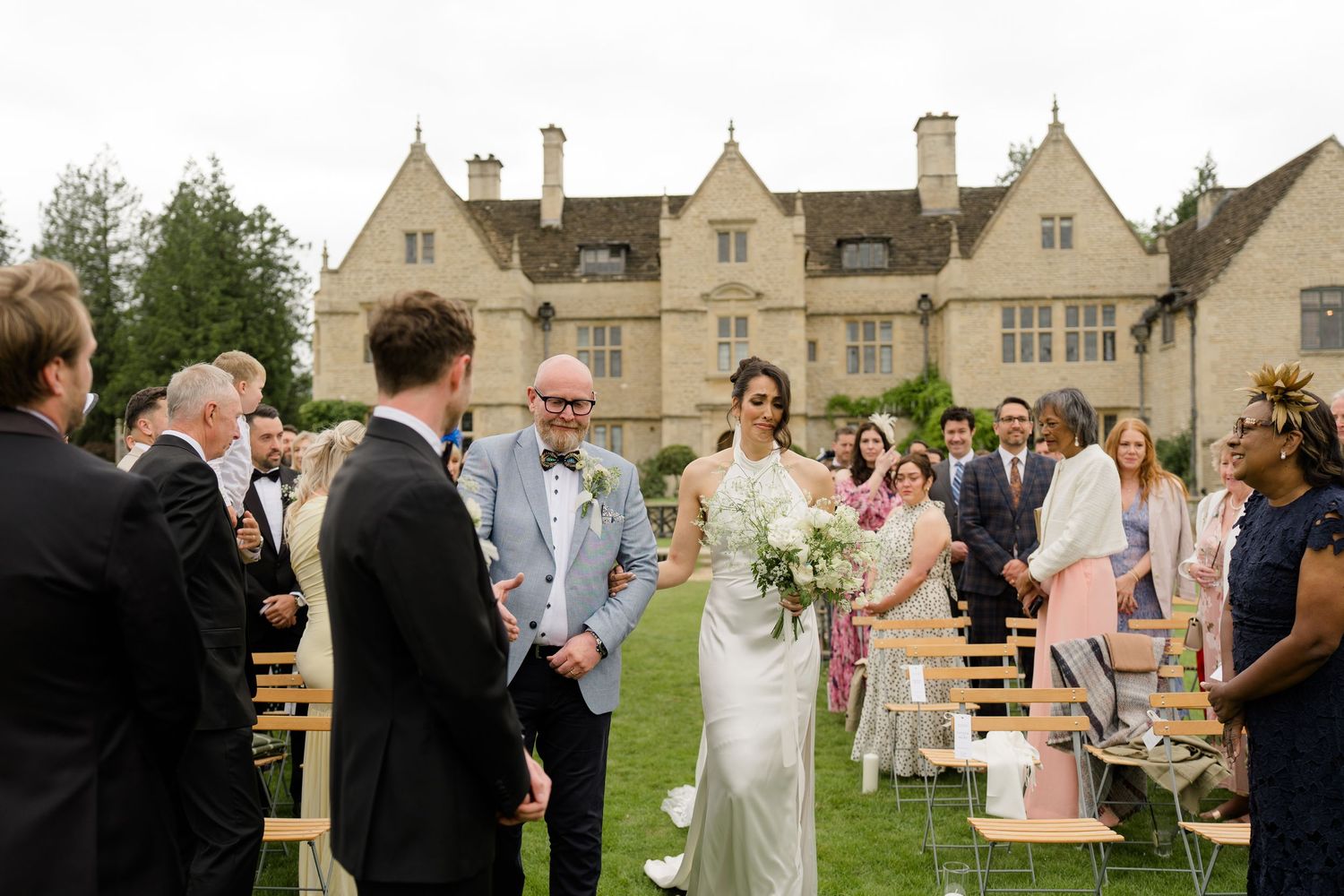 Bride in white gown walks down outdoor aisle with escort at a English manor wedding.