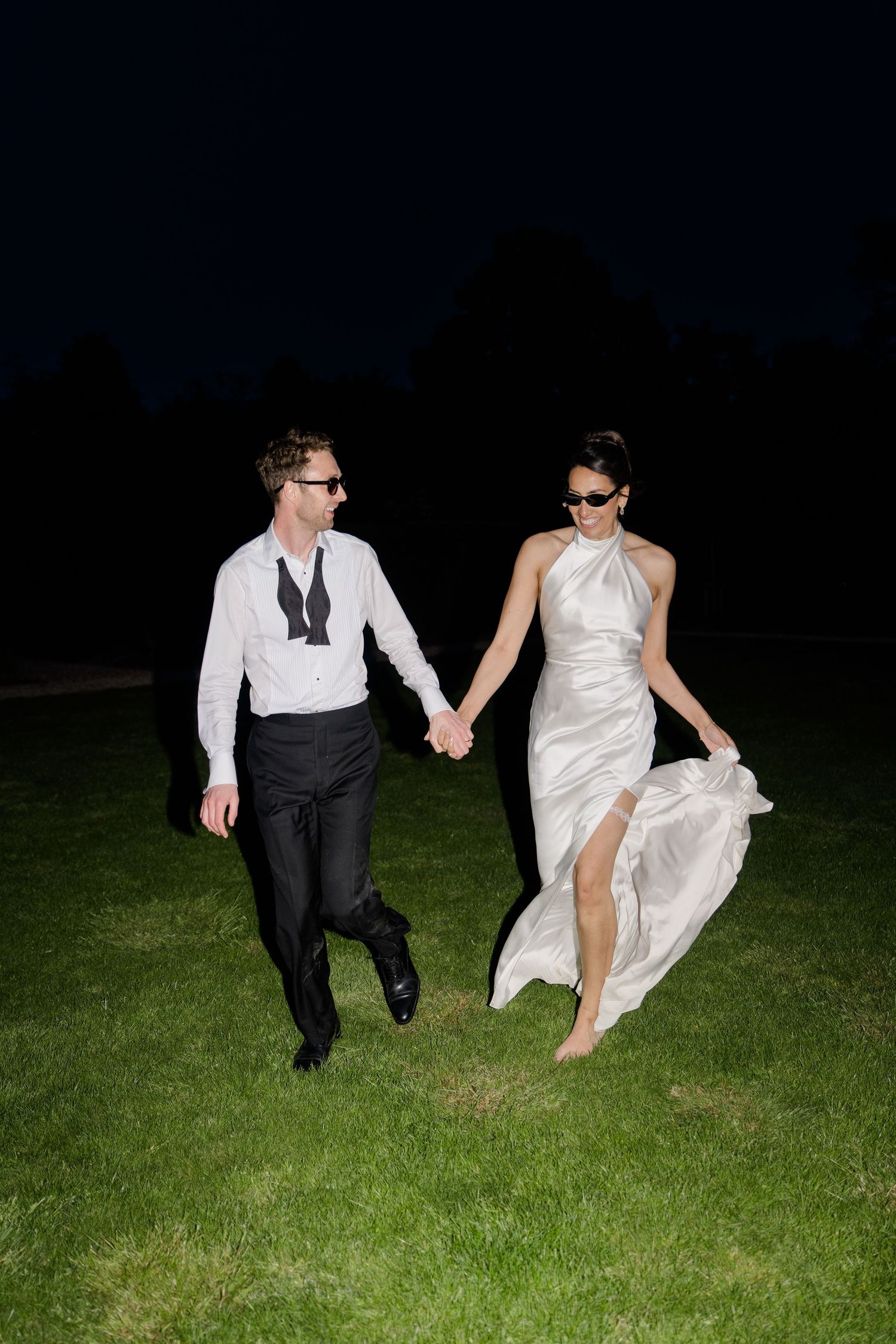 Bride and groom in sunglasses run hand in hand across dark lawn in nighttime wedding photo.