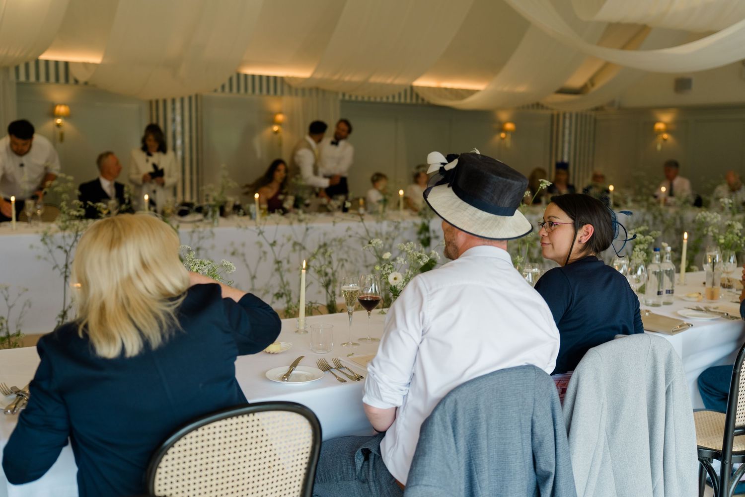 Wedding guests seated at elegantly decorated reception tables with greenery centerpieces and white draping above.