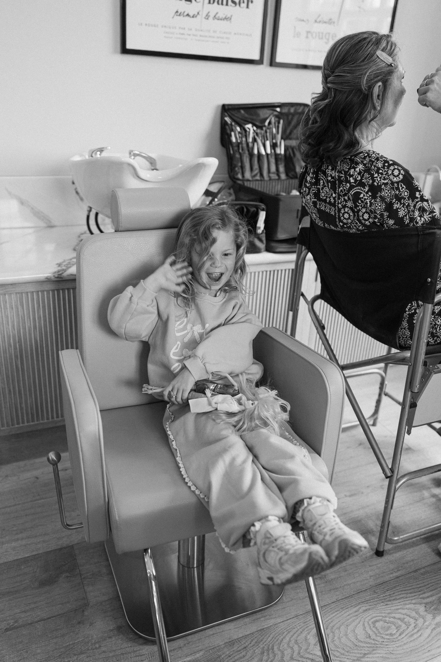 Black and white photo of a young girl laughing in a salon chair while a woman sits nearby at a styling station.