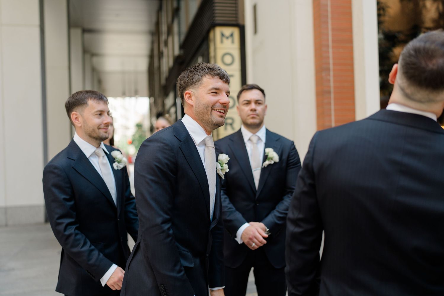 Smiling groom in dark suit with boutonniere stands with groomsmen before wedding ceremony outdoors.