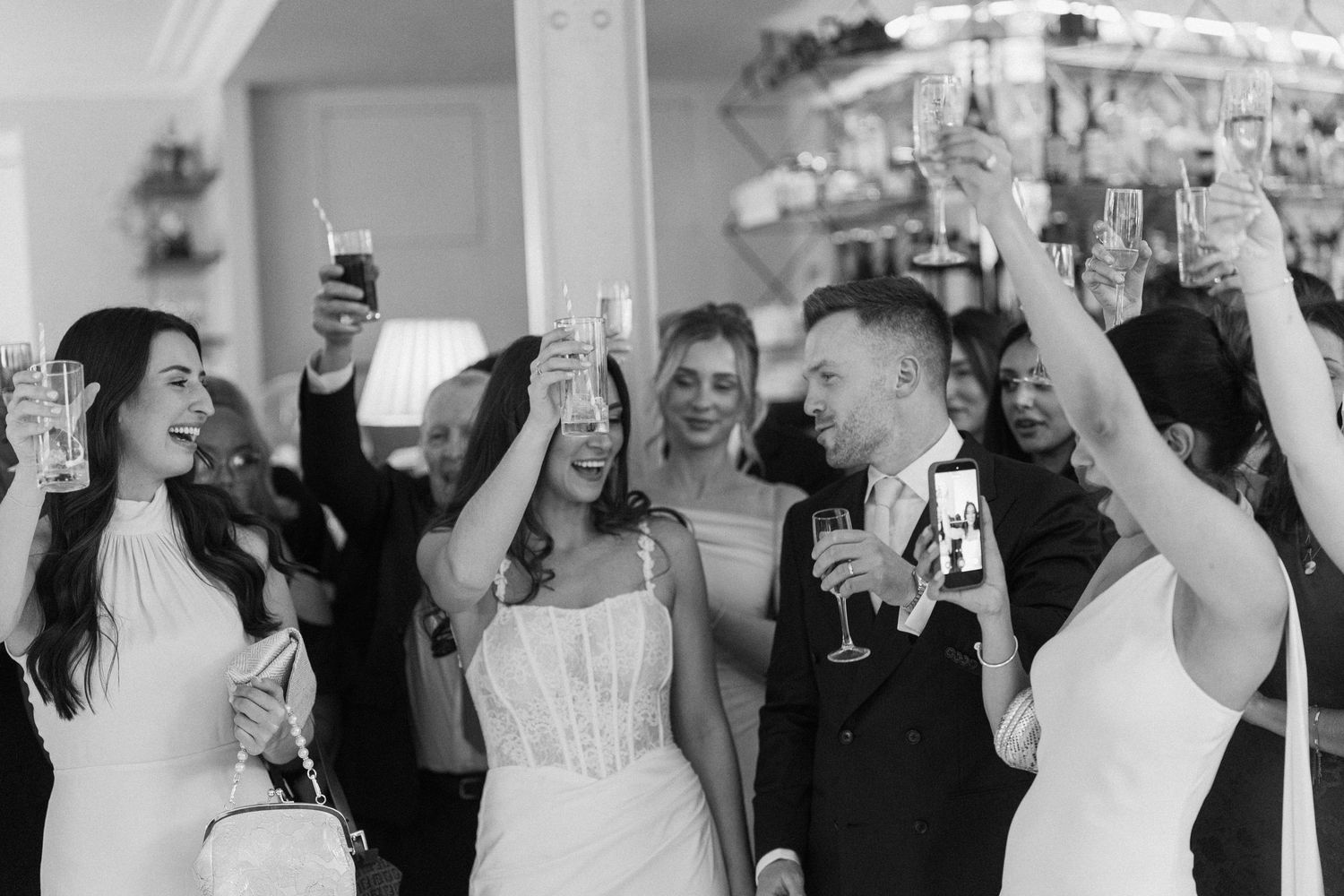Black and white photo of bride, groom and guests raising champagne glasses in a joyful wedding toast.