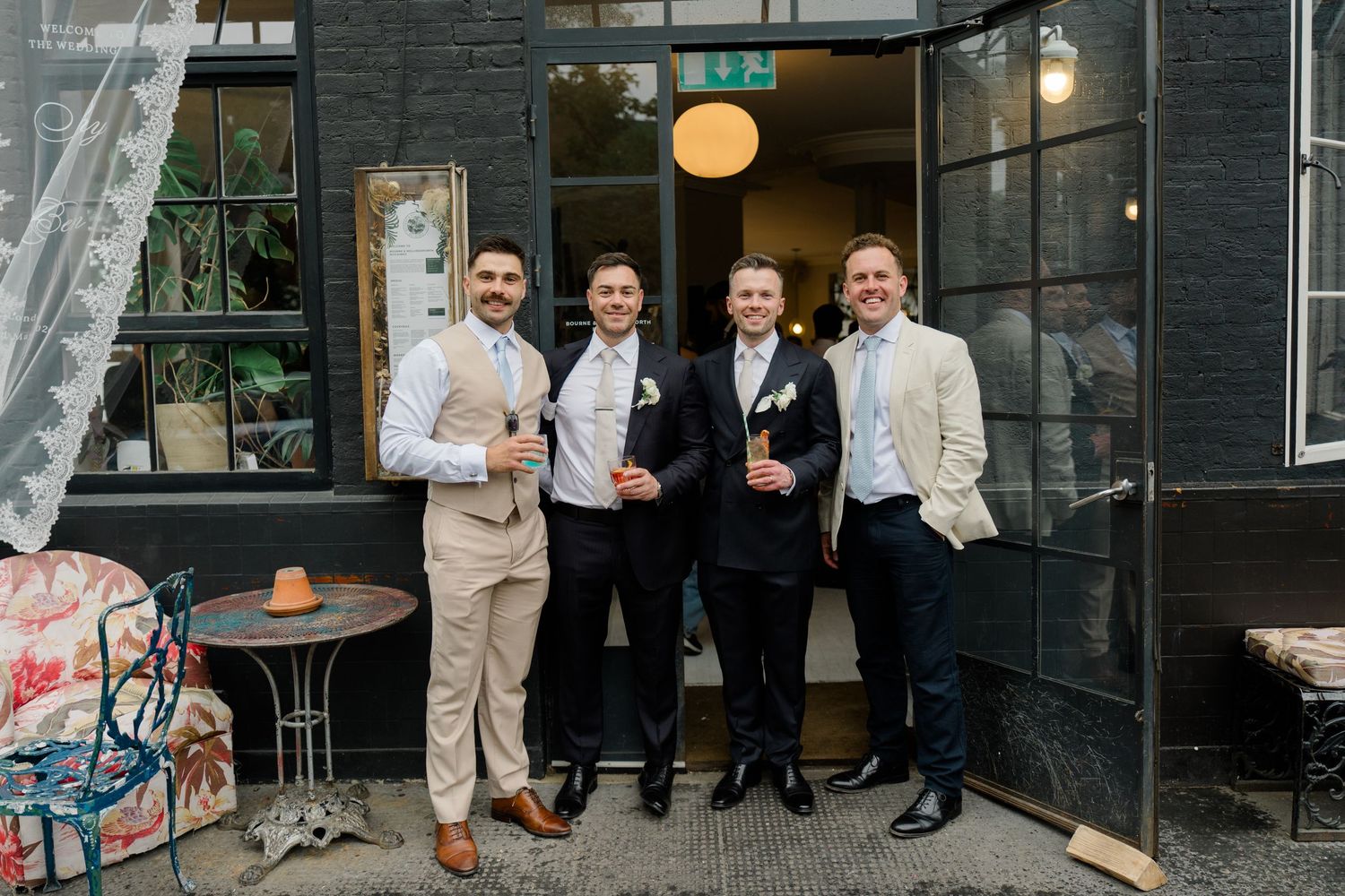 Four groomsmen smiling and holding drinks outside a dark-walled venue during an outdoor wedding reception.