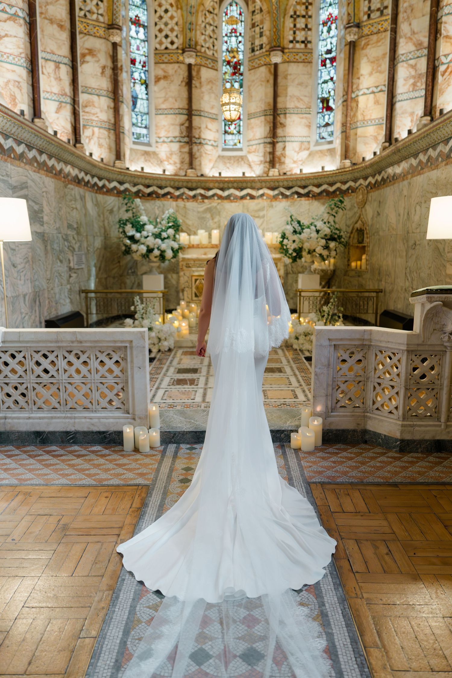 Bride in white gown and cathedral veil stands at chapel altar with stained glass windows and floral arrangements.