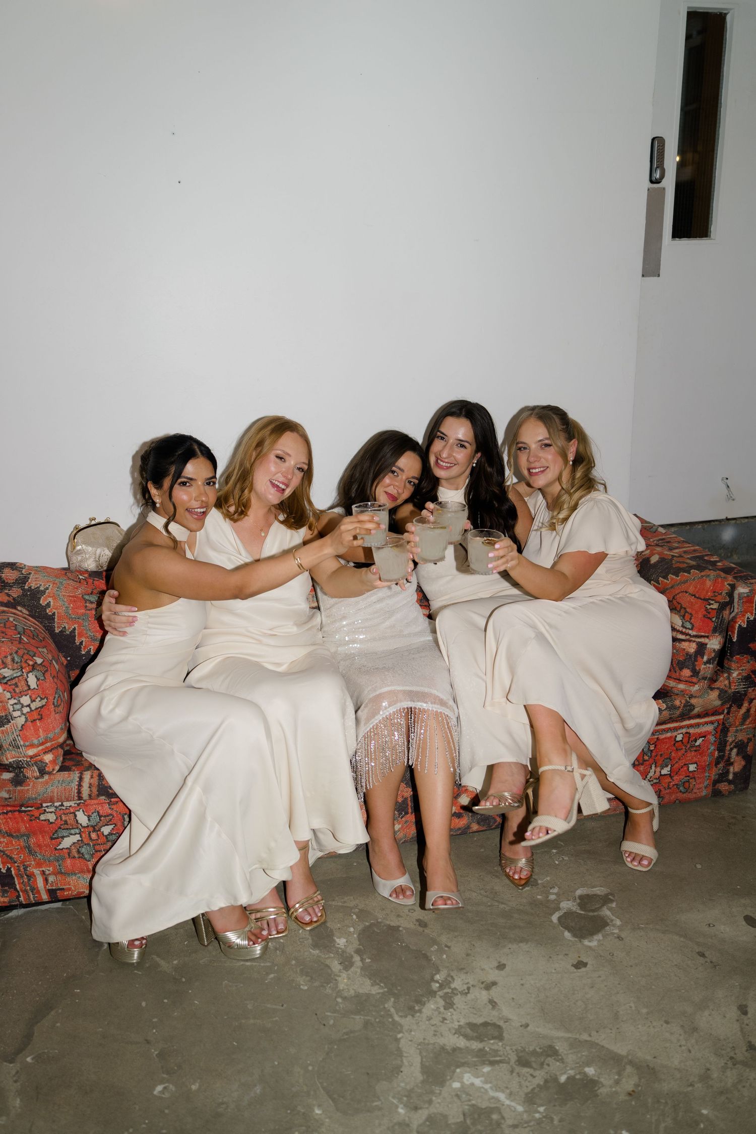 Five bridesmaids in white dresses toasting with drinks while seated on a floral sofa.