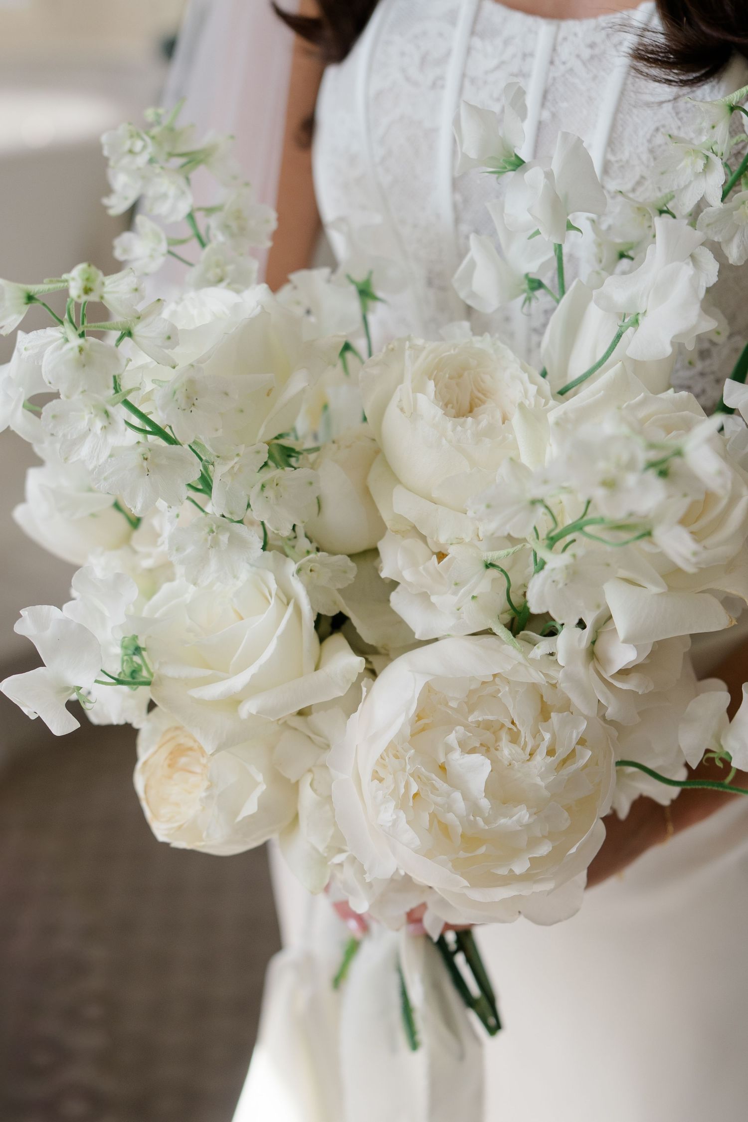 Bride holding lush white wedding bouquet of peonies and sweet peas with green accents.