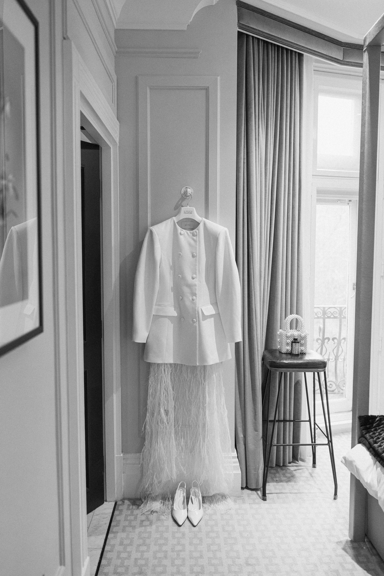 Elegant bridal outfit with white structured jacket and feathered skirt hanging in a Parisian hotel room with heels below.