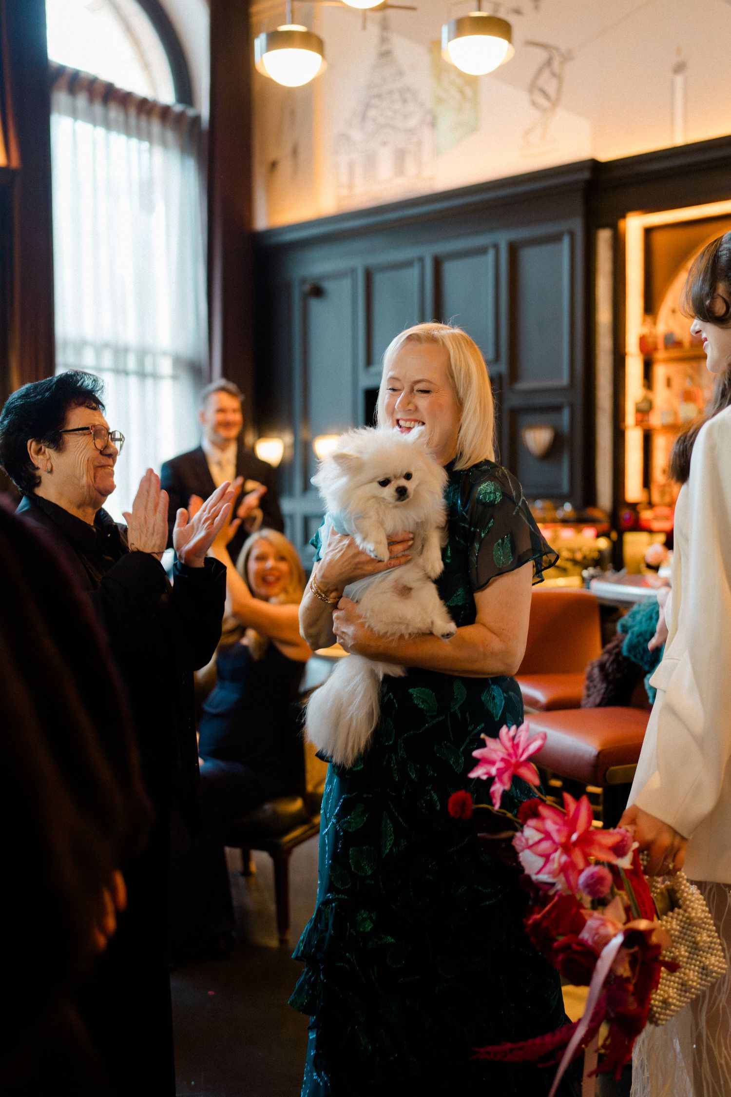Smiling woman in green dress holding white Pomeranian dog at elegant indoor wedding ceremony.