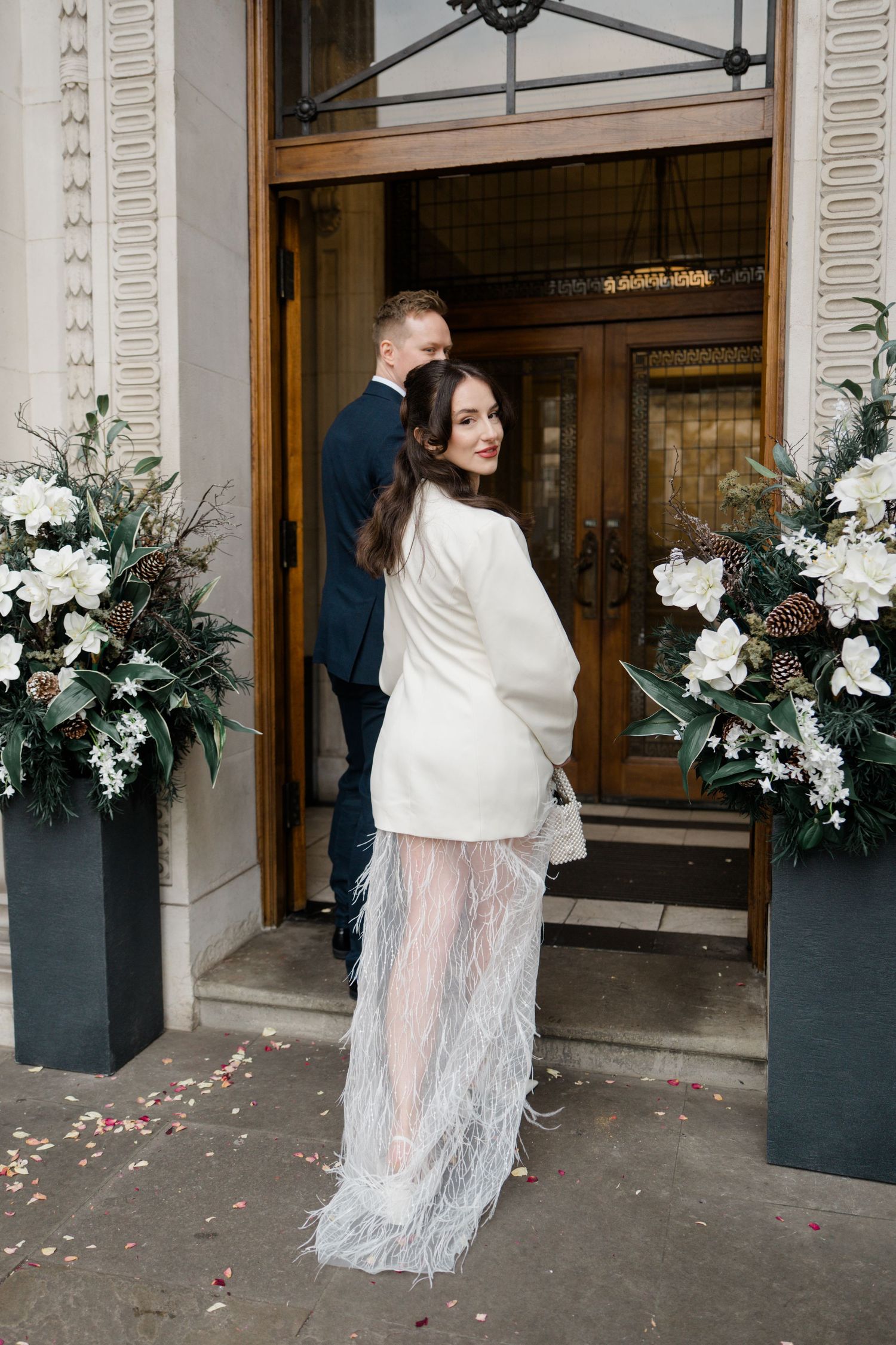 Bride in white blazer dress with feather hem turns to smile at entrance flanked by floral arrangements.