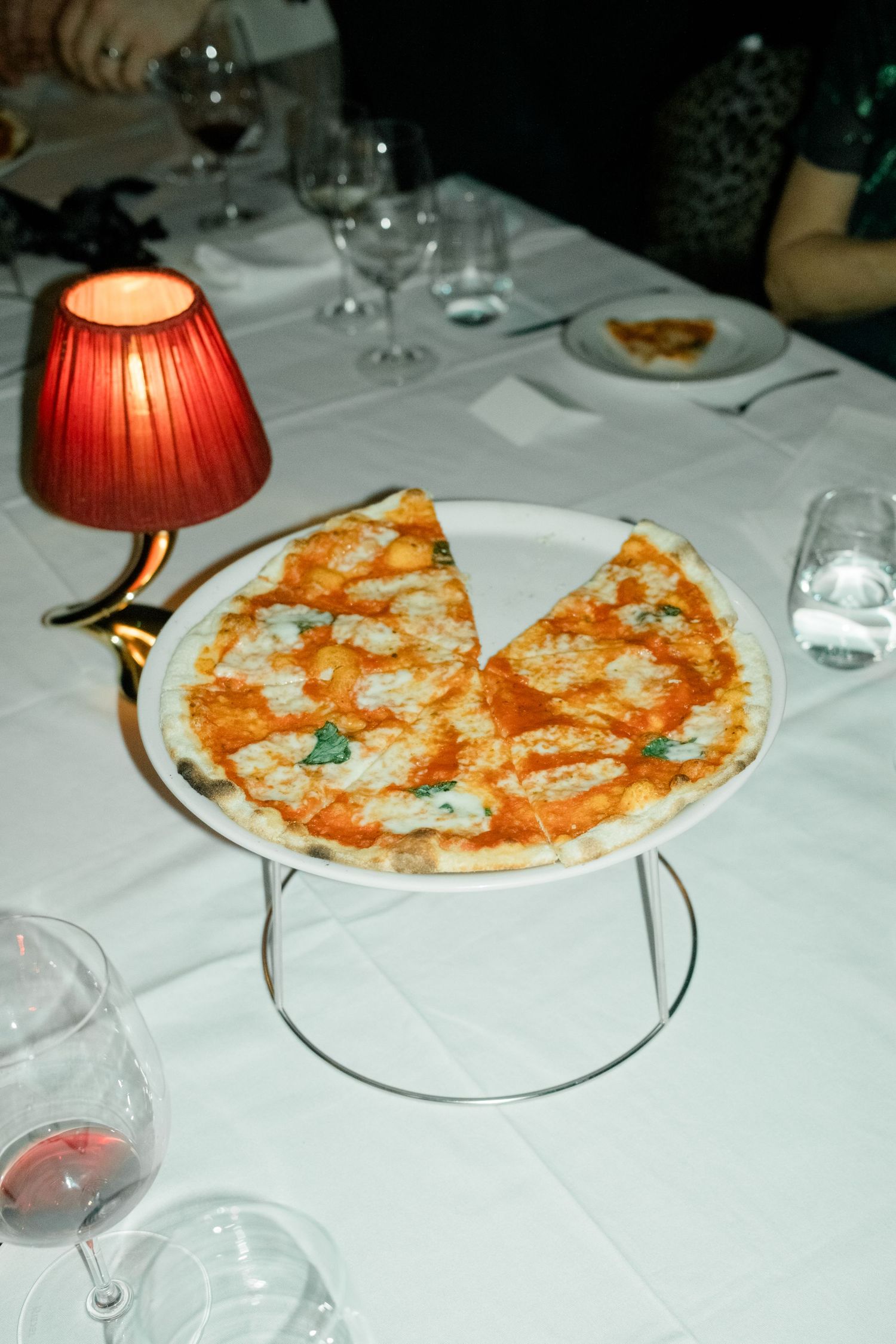 Margherita pizza served on a raised stand at an upscale restaurant table with wine glasses and a red lamp.