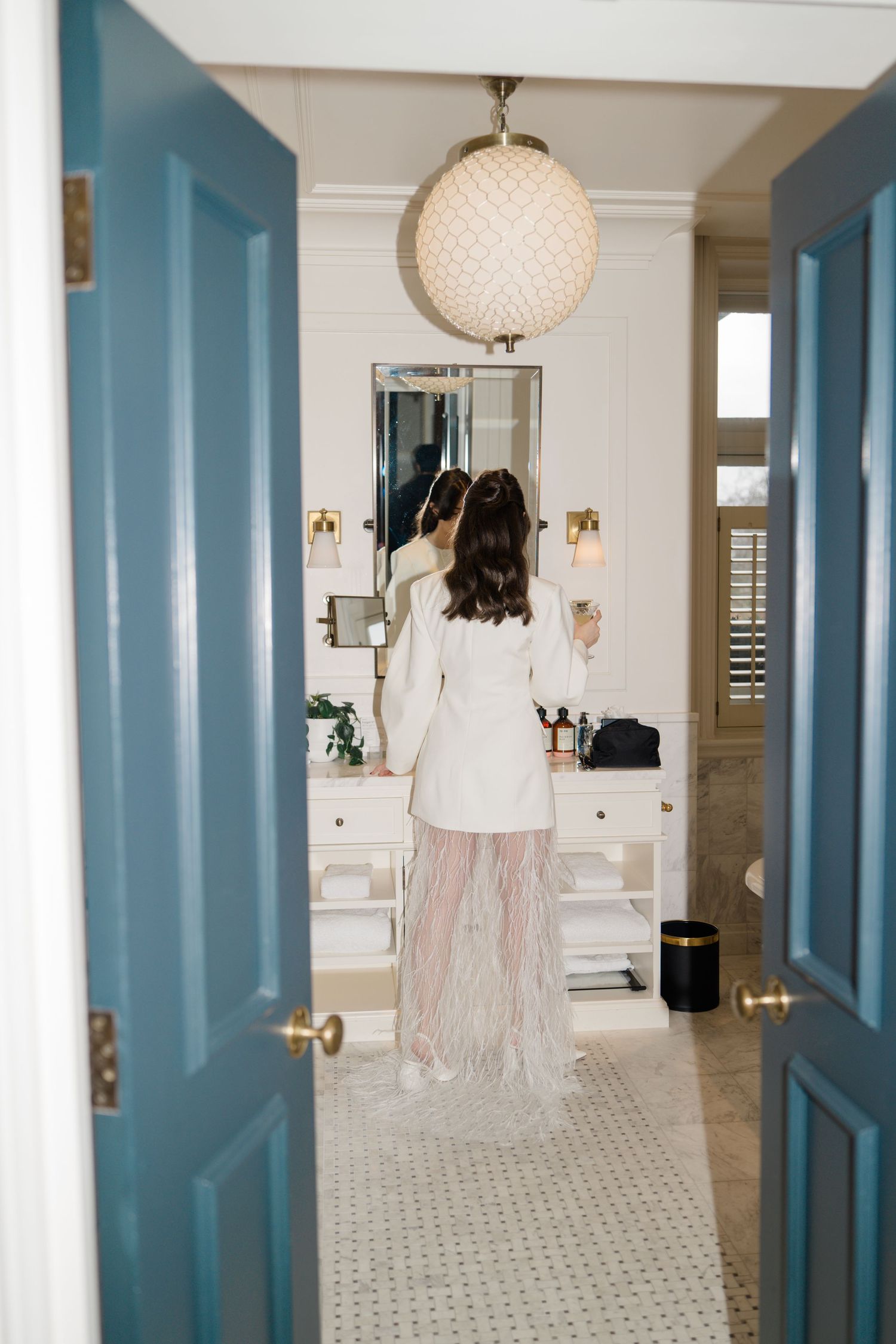 Bride in white blazer and feathered skirt admiring herself in a bathroom mirror through blue double doors.